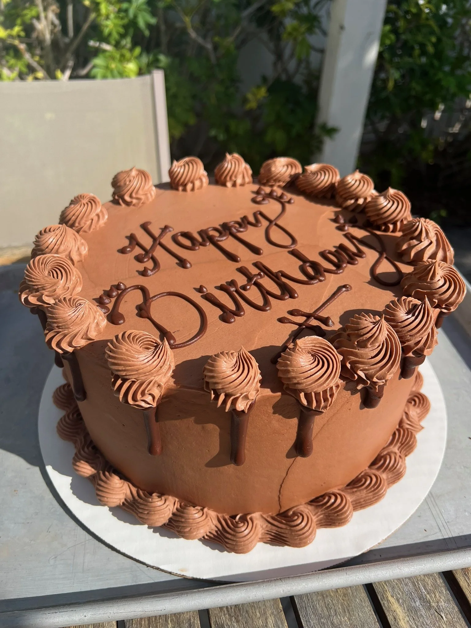 A chocolate cake on a white cake board that. Says happy birthday in a darker chocolate