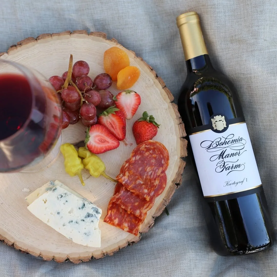 A cheese and charcuterie platter with grapes, strawberries, dried apricots, chili peppers, blue cheese, and sliced cured meat on a wooden slab, next to a bottle of red  Bohemia's wine and a glass of red wine.