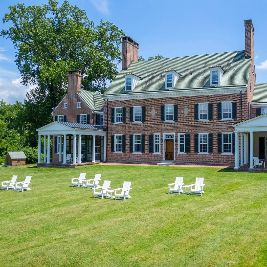 Bohemia Manor Farm.  A large brick house with multiple chimneys, green roof, and several windows. There are white chairs arranged on a well-maintained lawn in front of the house, with trees and a bright blue sky in the background.