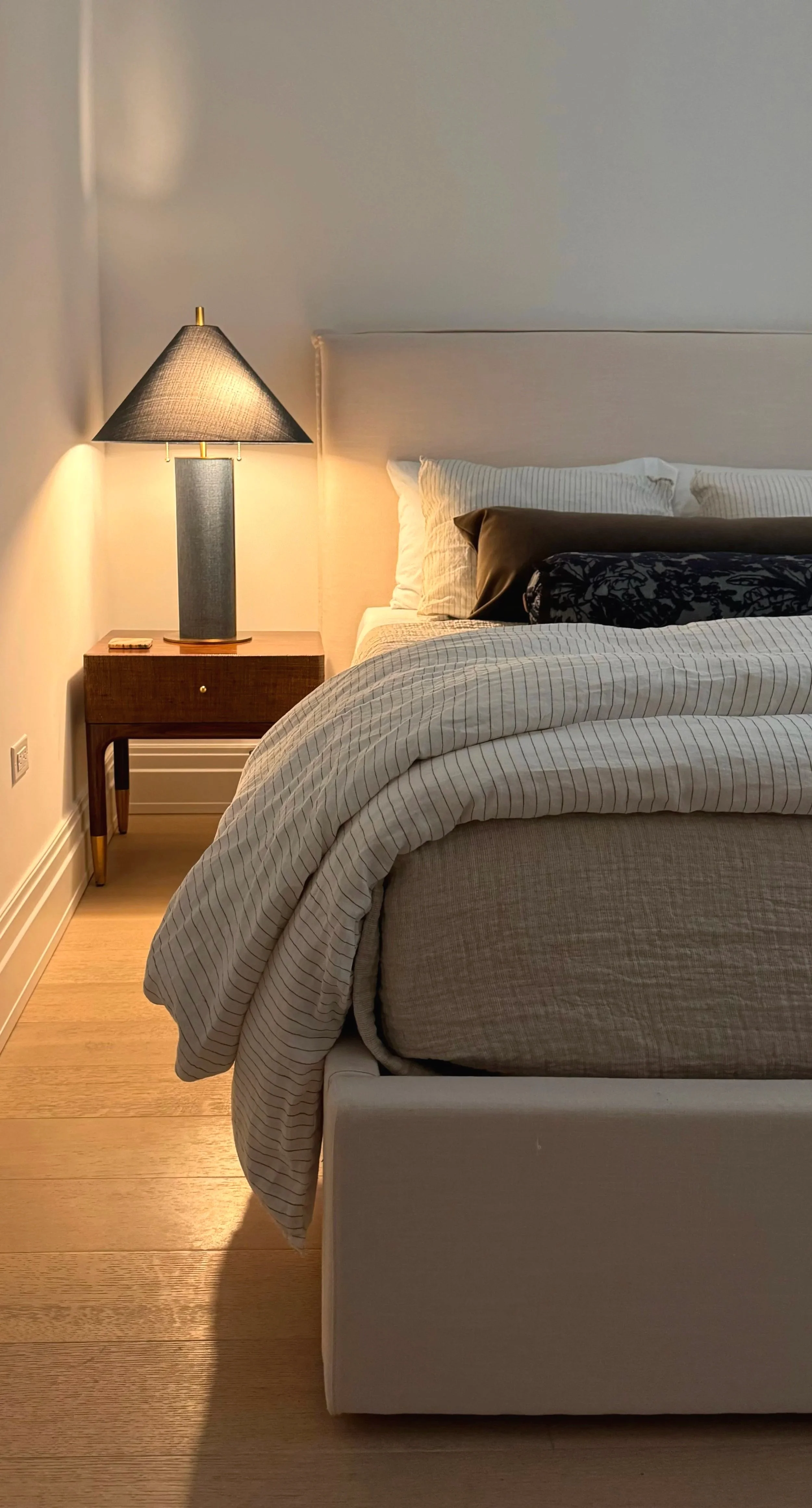 A neatly made bed with beige and black pillows in a modern bedroom, next to a wooden nightstand with a black lampshade and a small item on top.
