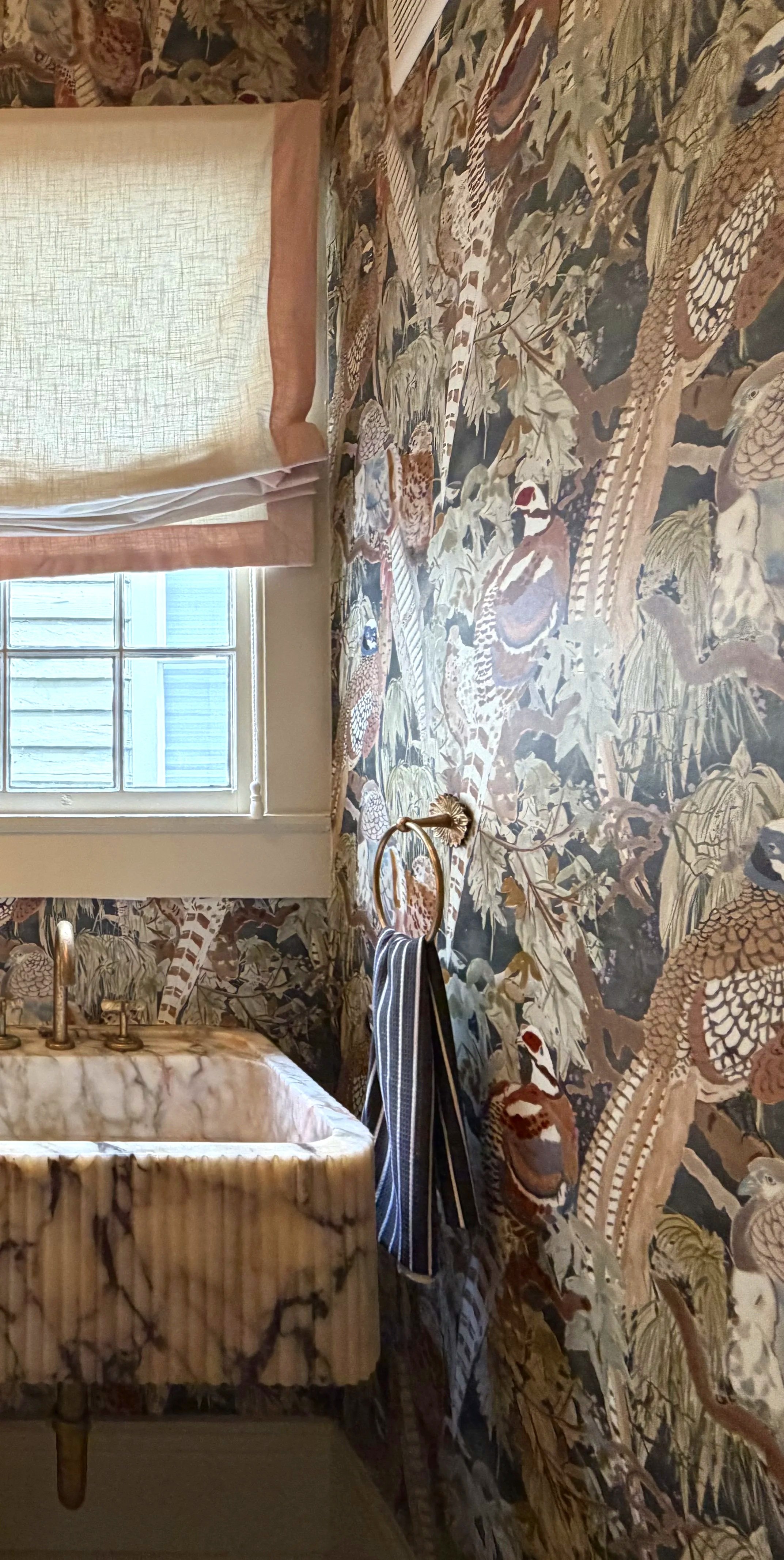 A room with jungle-themed wallpaper featuring various animals and plants, a window with a beige shade, a marble-top dresser, and a striped bag hanging on a gold ring hook.