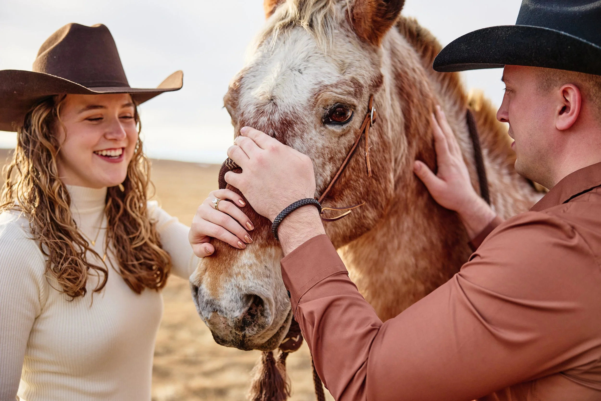 A woman and a man, both wearing cowboy hats, are petting and interacting with a light brown horse against a background of an open field.