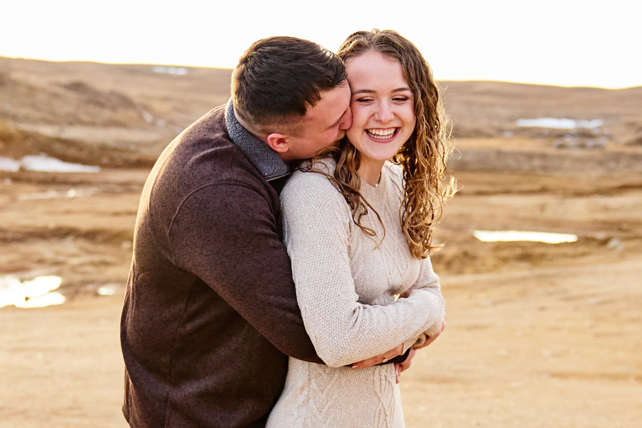 A couple in warm clothing embracing outdoors in a landscape with rolling hills and patches of snow, at sunset or sunrise.