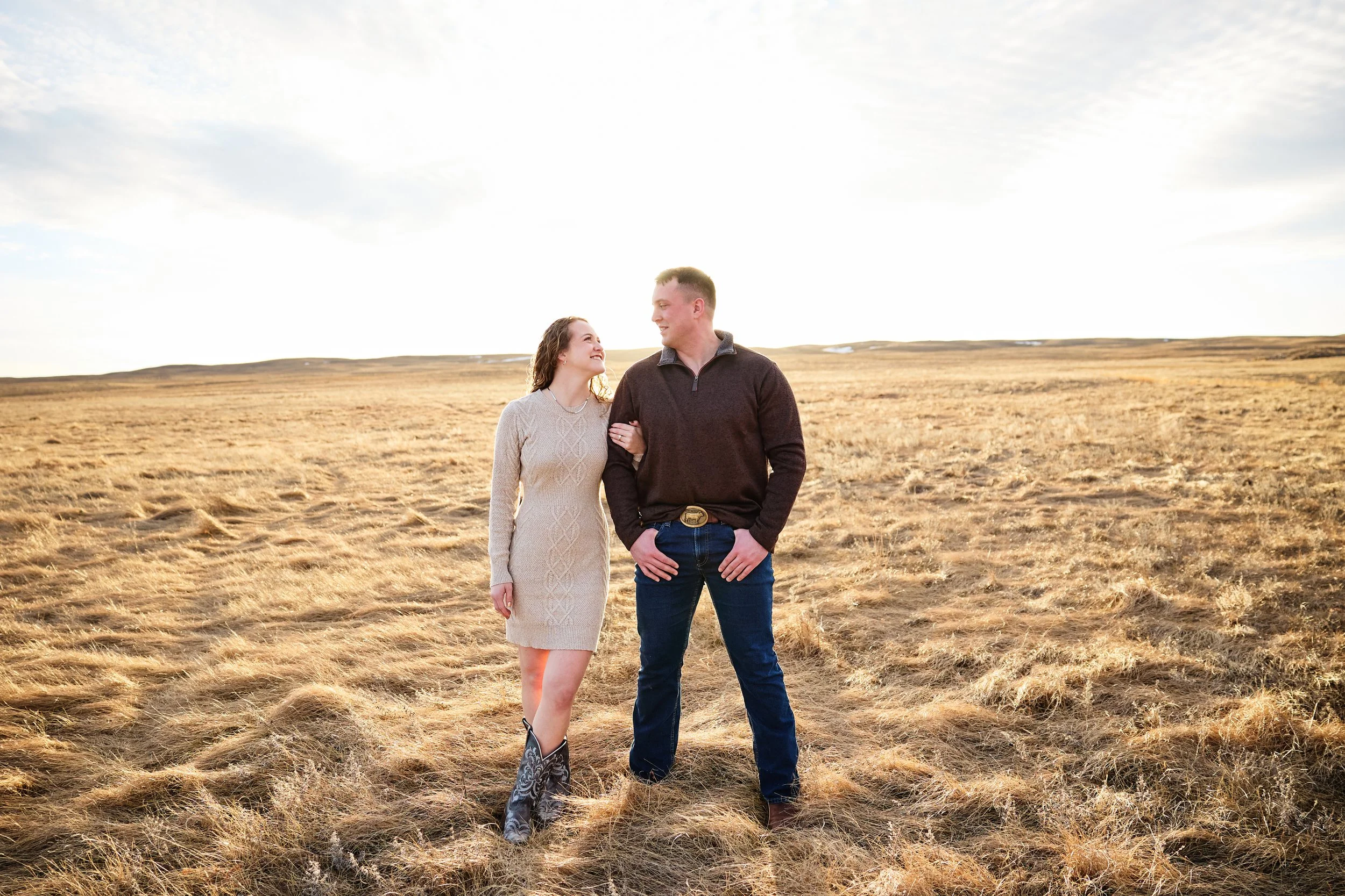 A couple standing in a field of dry grass during sunset, smiling and looking at each other, with a bright sky overhead.