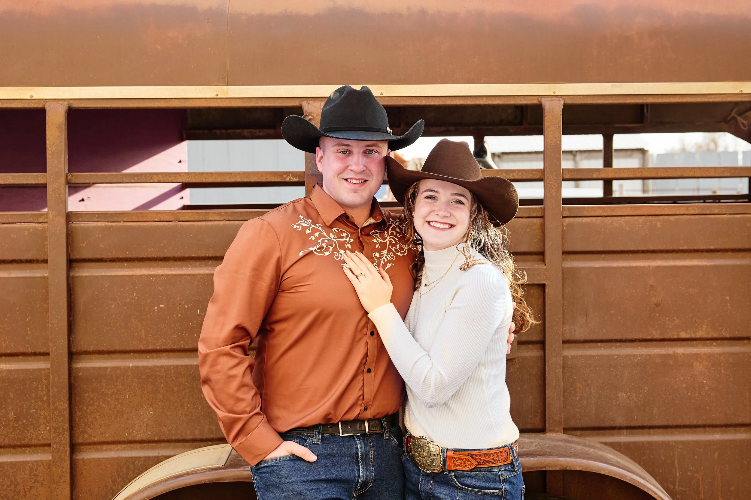 A young man and woman in western attire smiling and posing together in front of a rusted cattle trailer. The man is wearing a black cowboy hat, brown button-up shirt with embroidery, and jeans. The woman is wearing a brown cowboy hat, white sweater, 