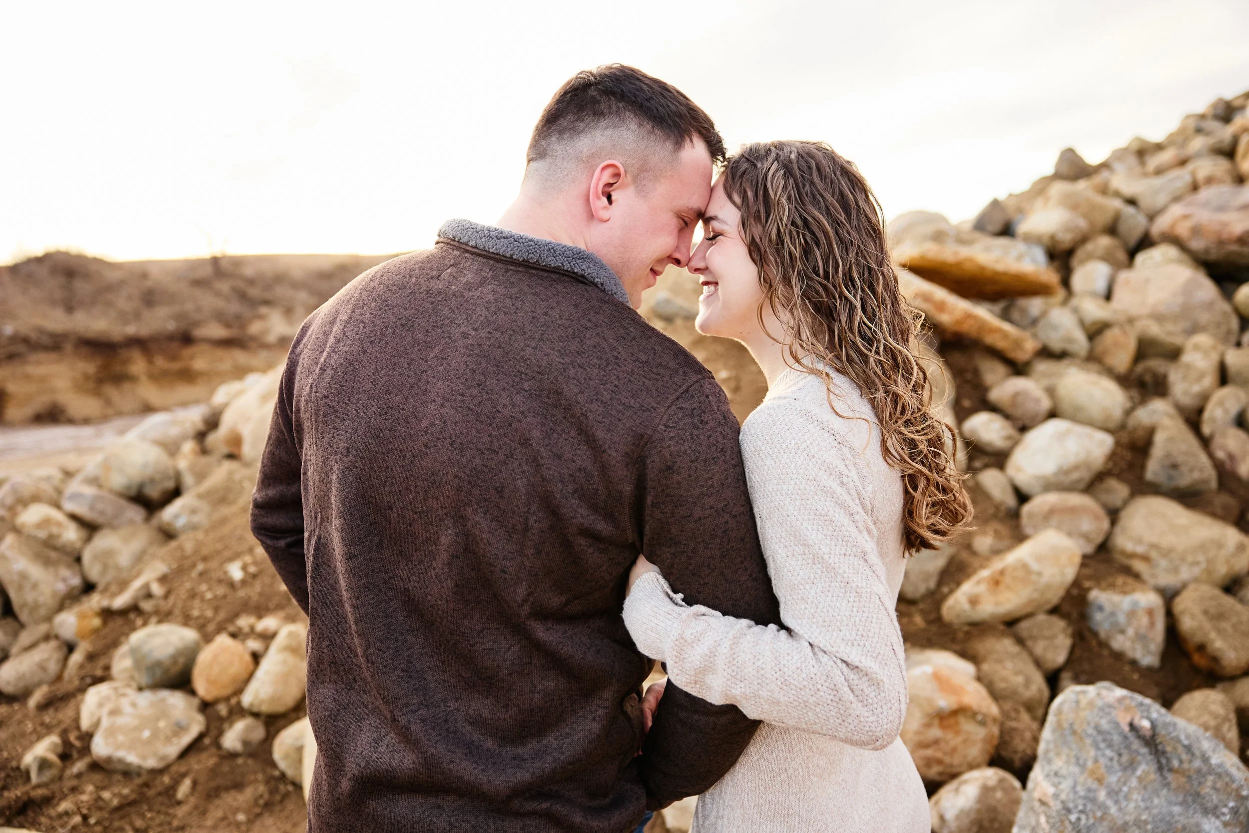 A young couple standing close, touching foreheads and smiling in a rocky outdoor setting with a soft sky background.