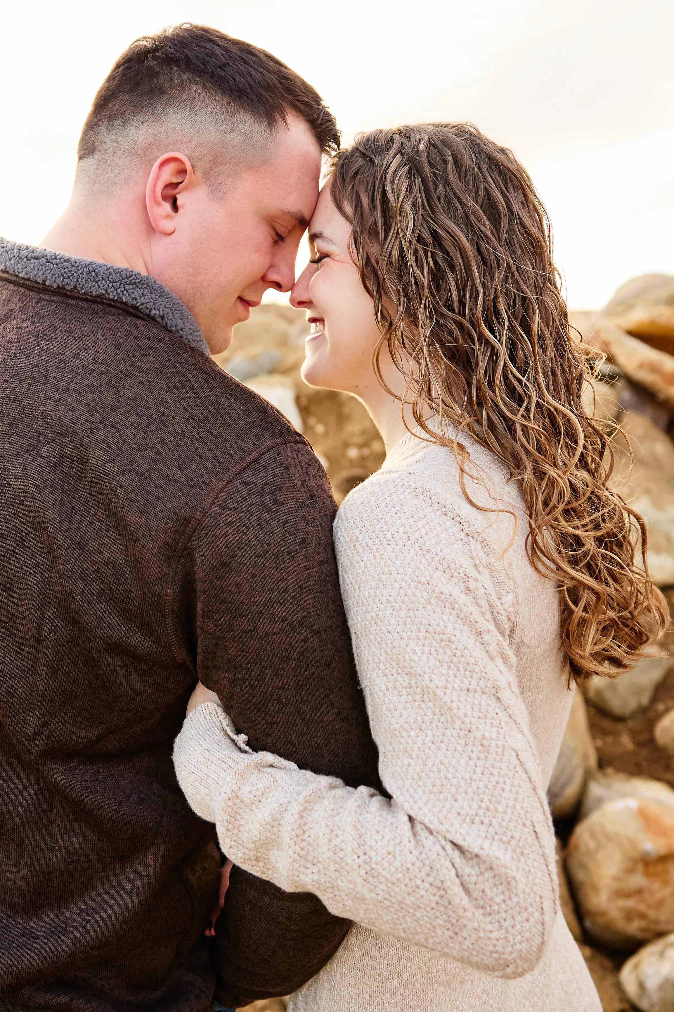 A couple standing close together, touching foreheads, smiling, outdoors near rocks at sunset.