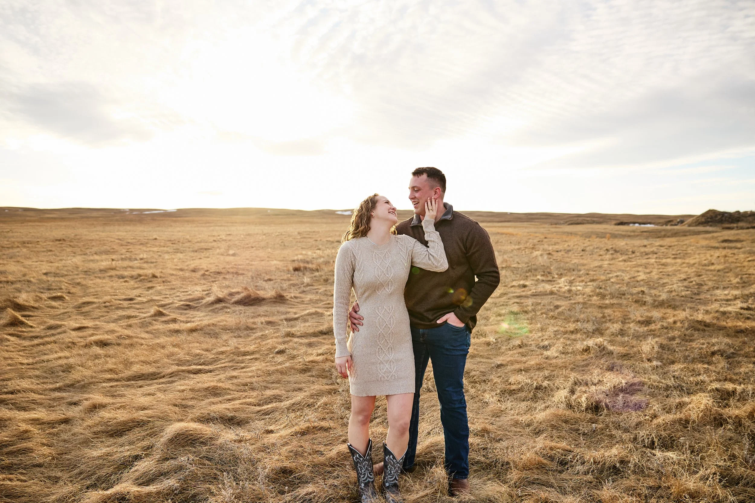 A couple standing in a grassy field at sunset, smiling and looking at each other, with open sky and clouds in the background.