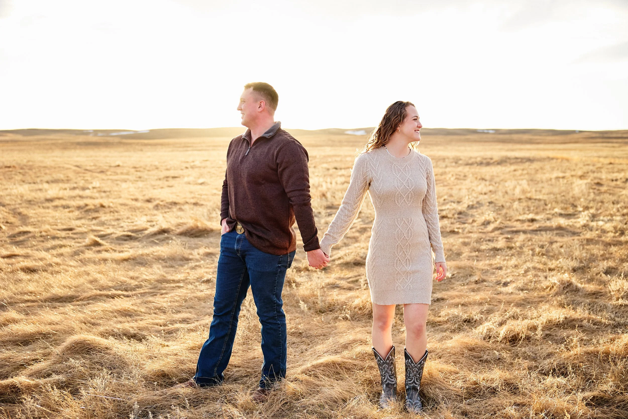 A man and woman holding hands and smiling, standing in a grassy field during sunset.