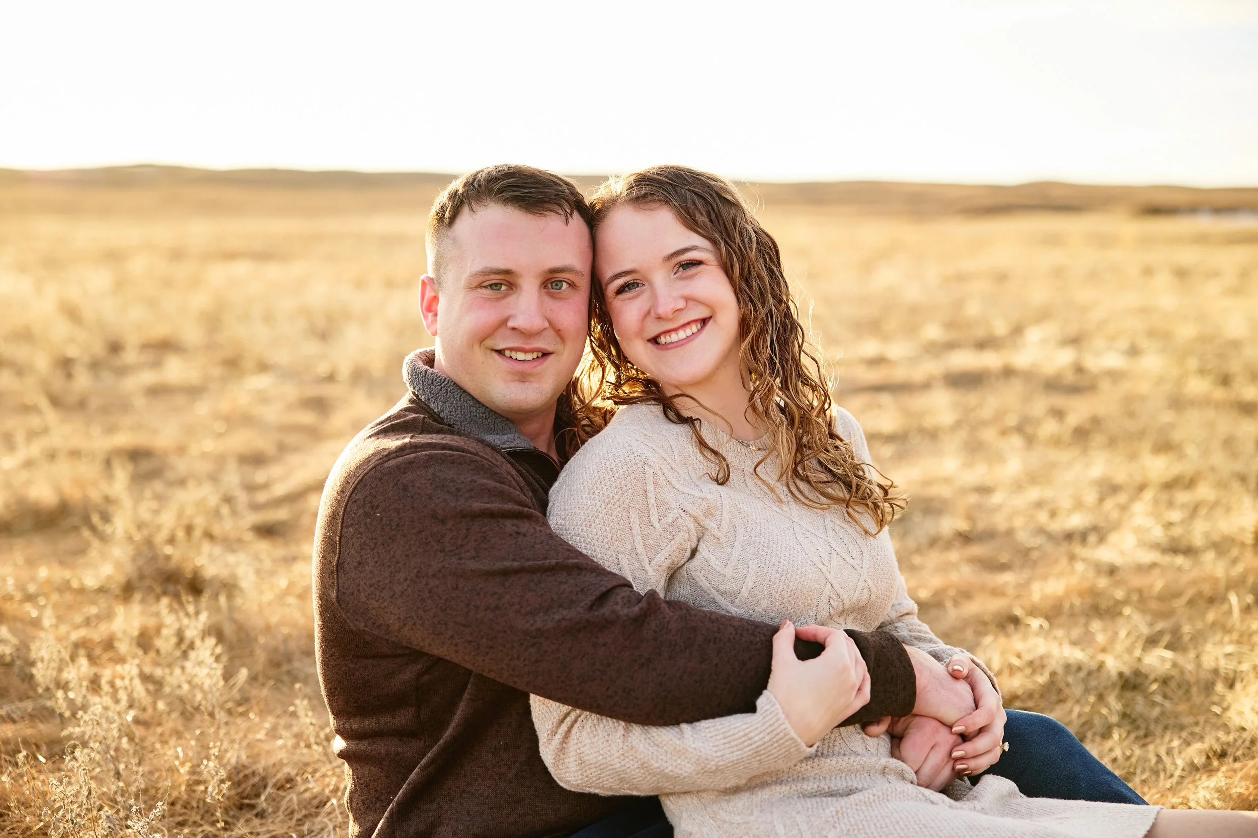 A happy couple sitting close together in a field during sunset, smiling at the camera.