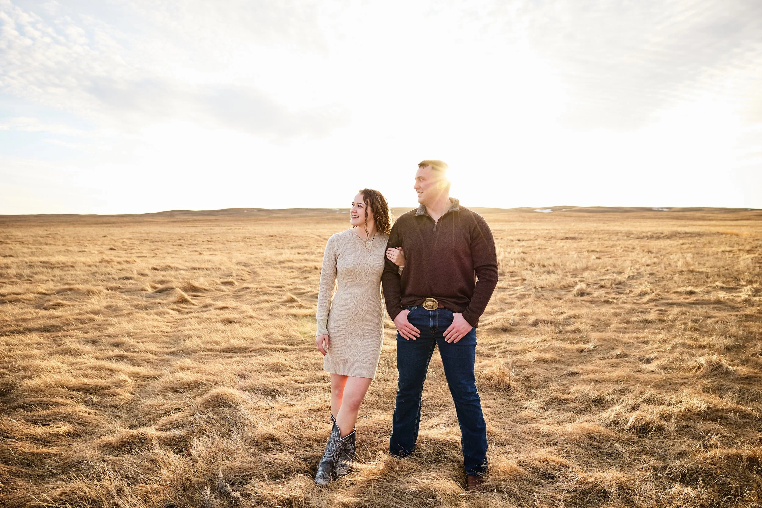 A couple walking arm-in-arm through a grassy field at sunset, smiling, with the sun behind them and the sky partly cloudy.