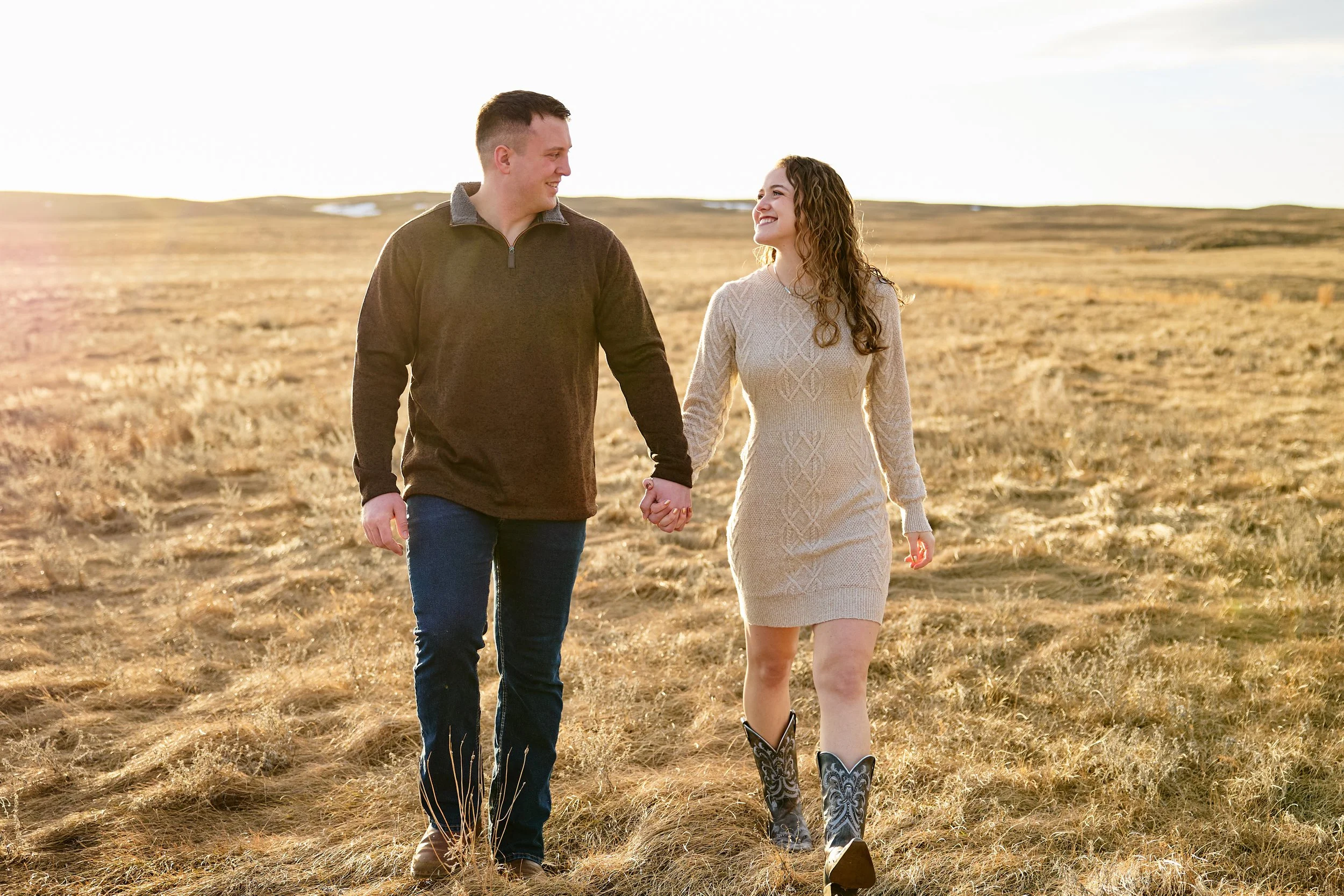 A young couple holding hands and walking through a grassy field during sunset, smiling at each other.