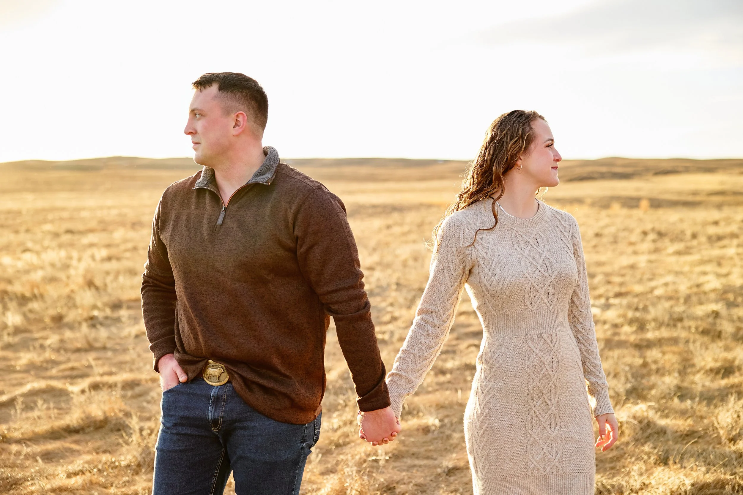 A couple holding hands and facing away from each other in an open field during sunset.