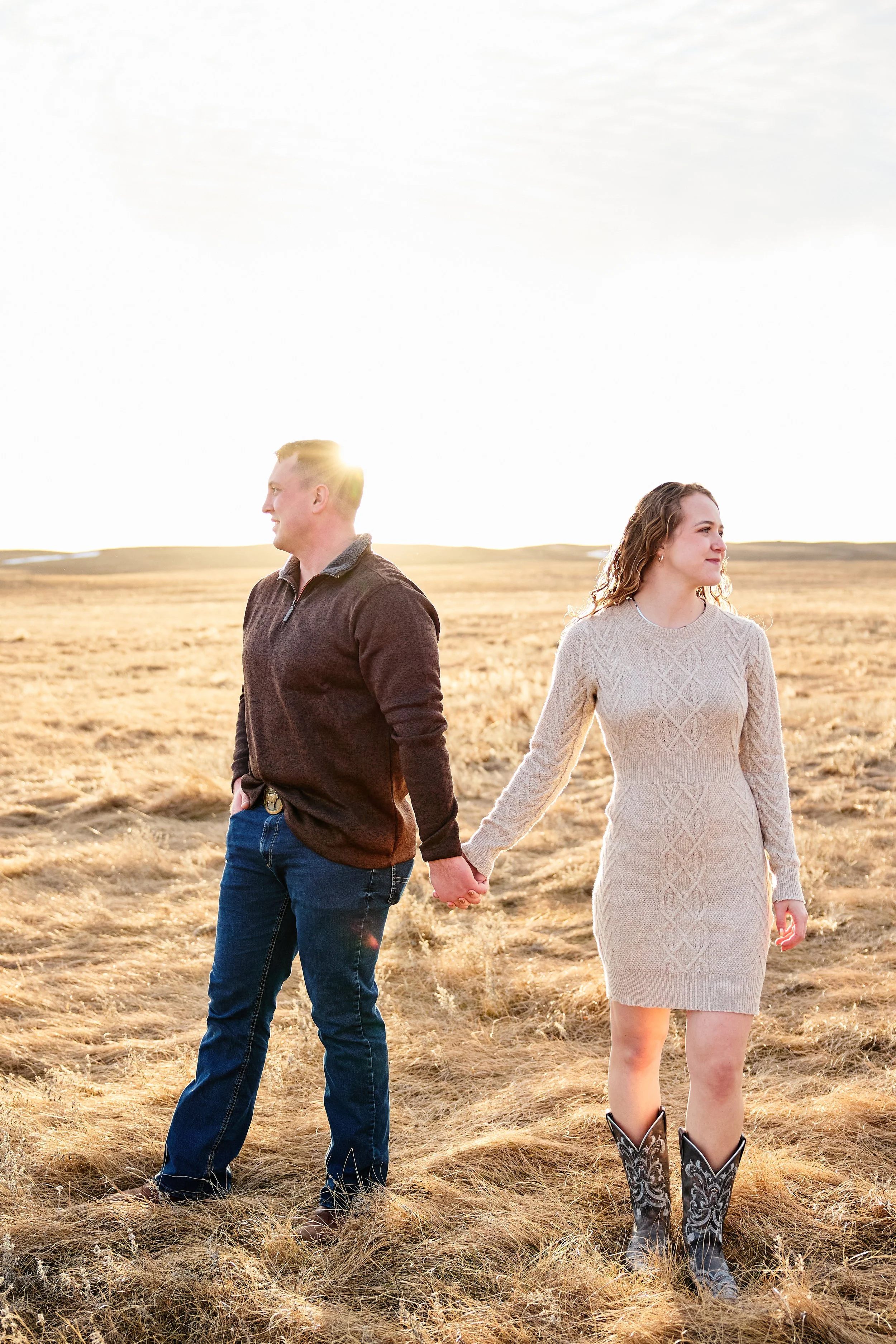 A man and woman holding hands in a field during sunset, facing opposite directions.