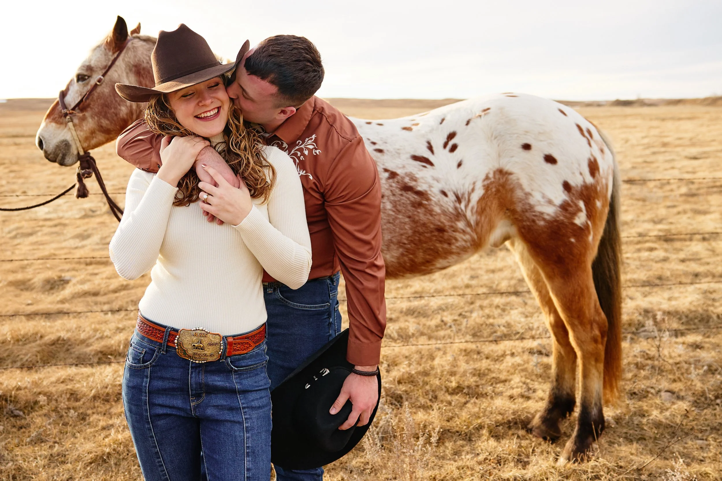 A couple sharing a joyful moment with a smiling woman wearing a white long-sleeve shirt and a cowboy hat, and a man in a brown shirt, hugging near a white and brown spotted horse in a grassy field with a cloudy sky.