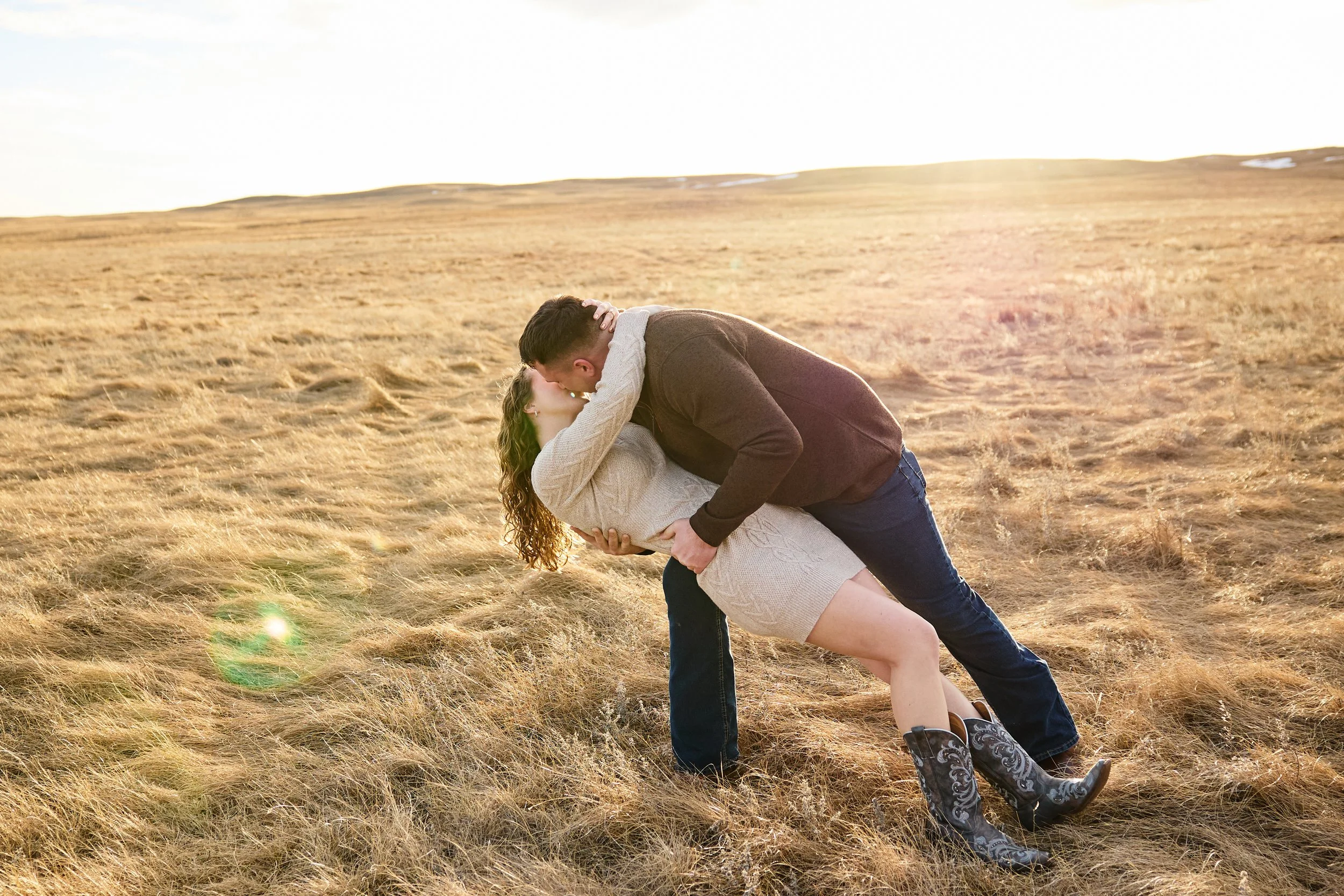 A man and woman sharing a kiss in a grassy field at sunset, with the man dipping the woman.