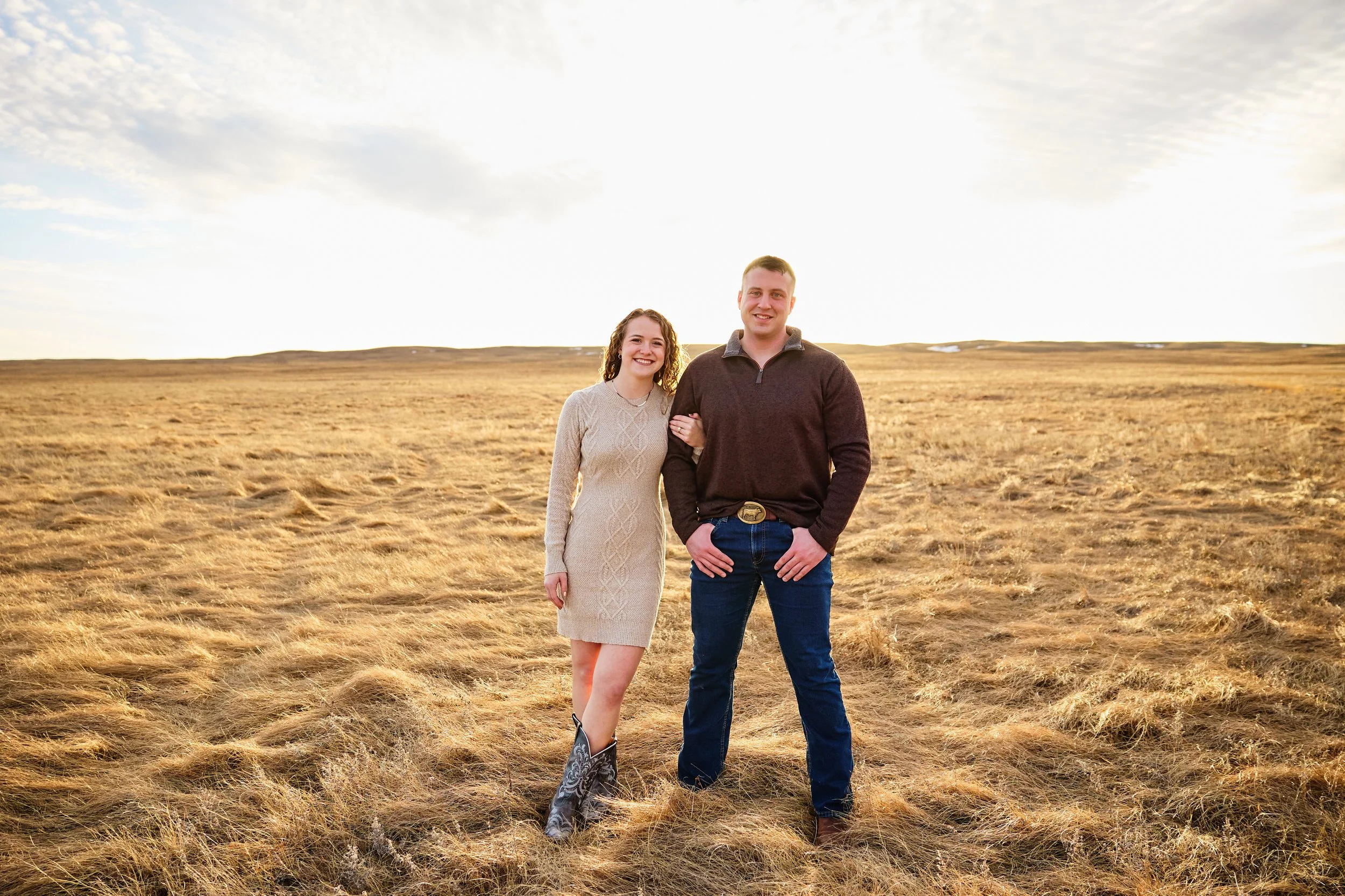 A young couple standing arm in arm in a grassy field during sunset, smiling at the camera.