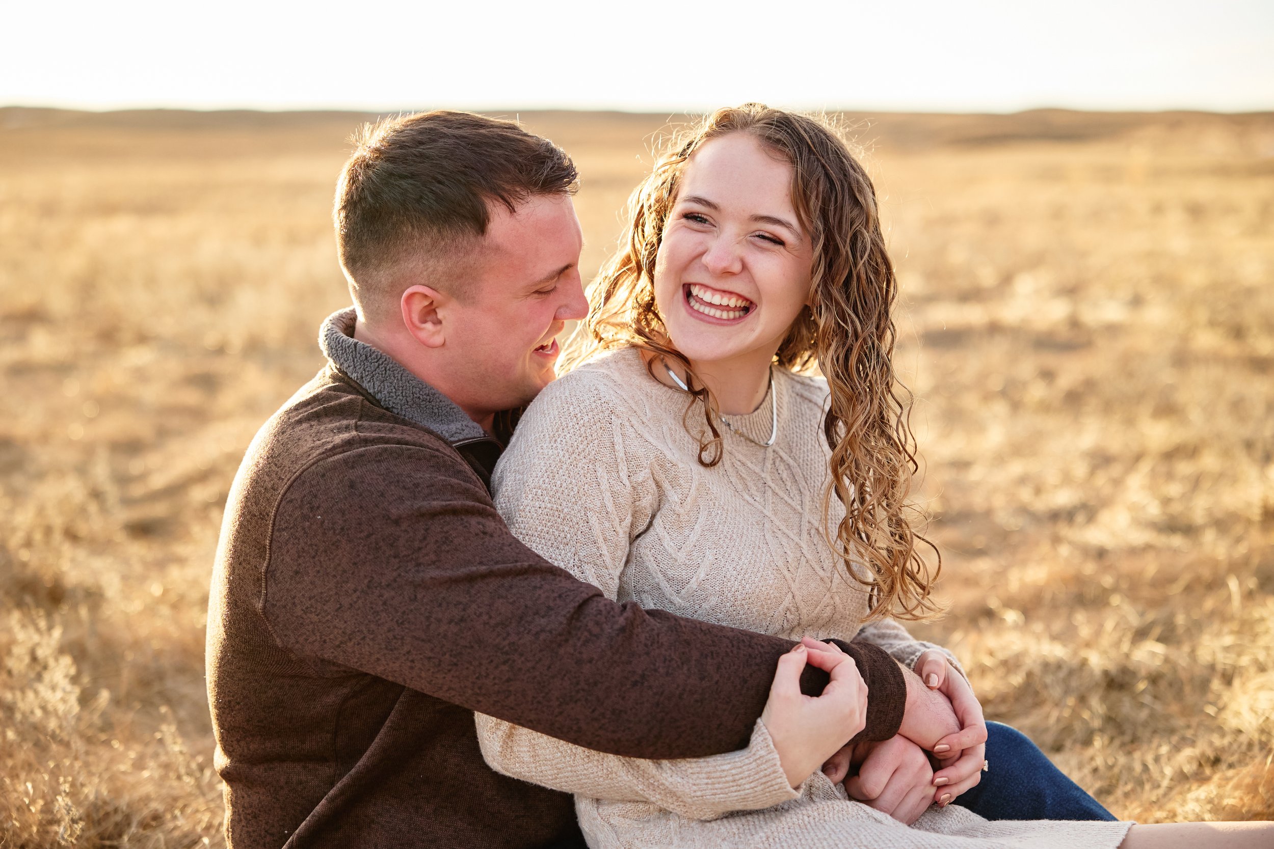 A happy couple sitting outdoors in a field, embracing and smiling at each other during sunset.