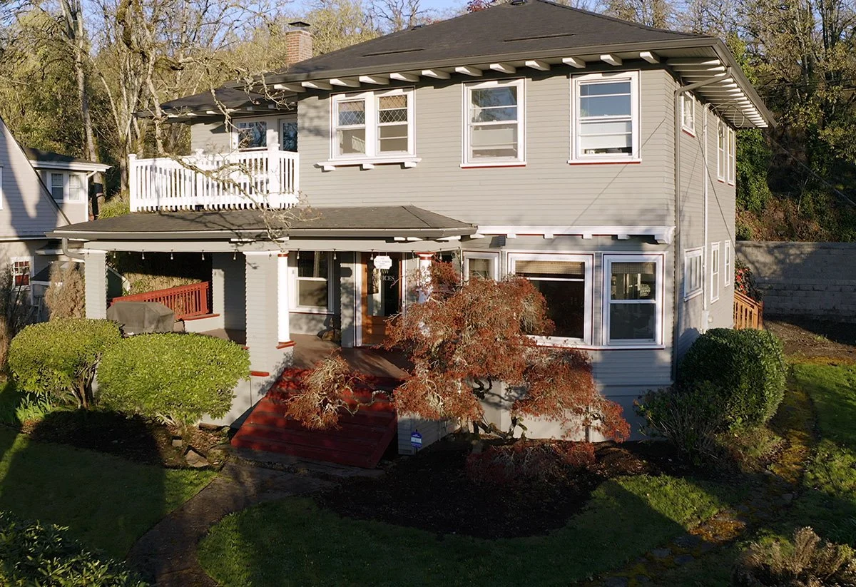 A two-story house with gray siding, white trim, multiple windows, and a small front porch surrounded by bushes and trees.