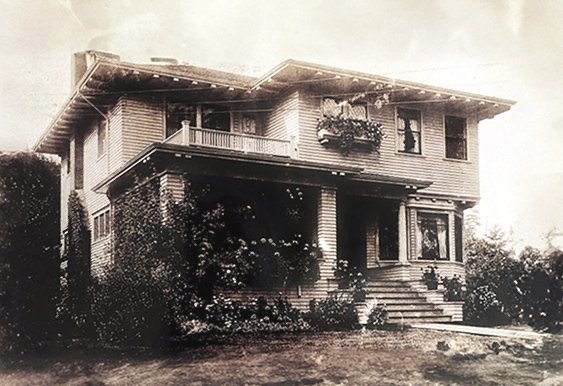 A black and white photo of a two-story house with a porch, stairs leading to the front door, and a garden area with bushes and plants.