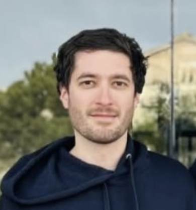 Joven con barba y cabello oscuro, usando sudadera azul, de pie al aire libre con edificios y árboles en el fondo.