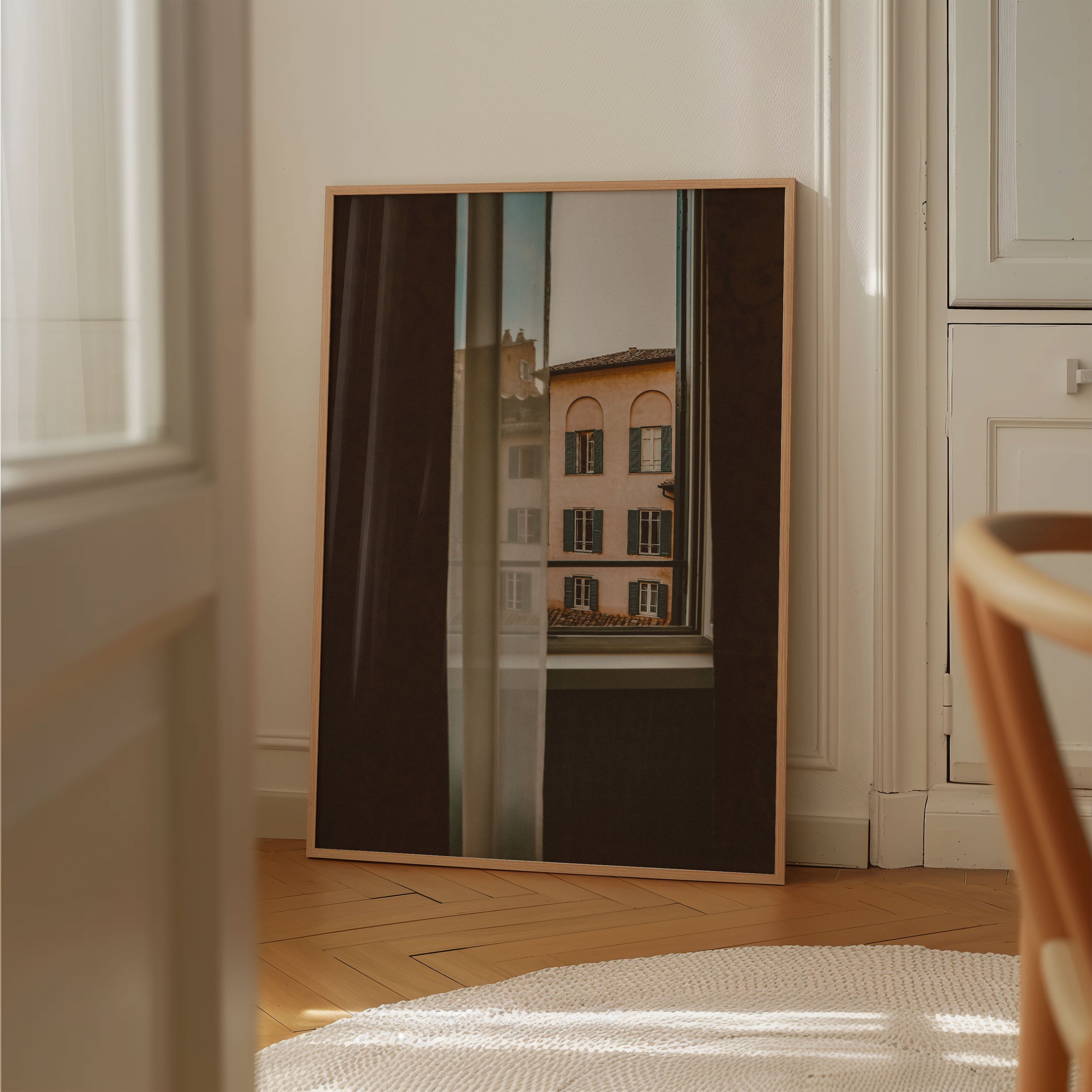 A framed mirror leaning against a wall in a room with white walls and wooden flooring. The mirror reflects a window showing a building with multiple windows and green shutters.