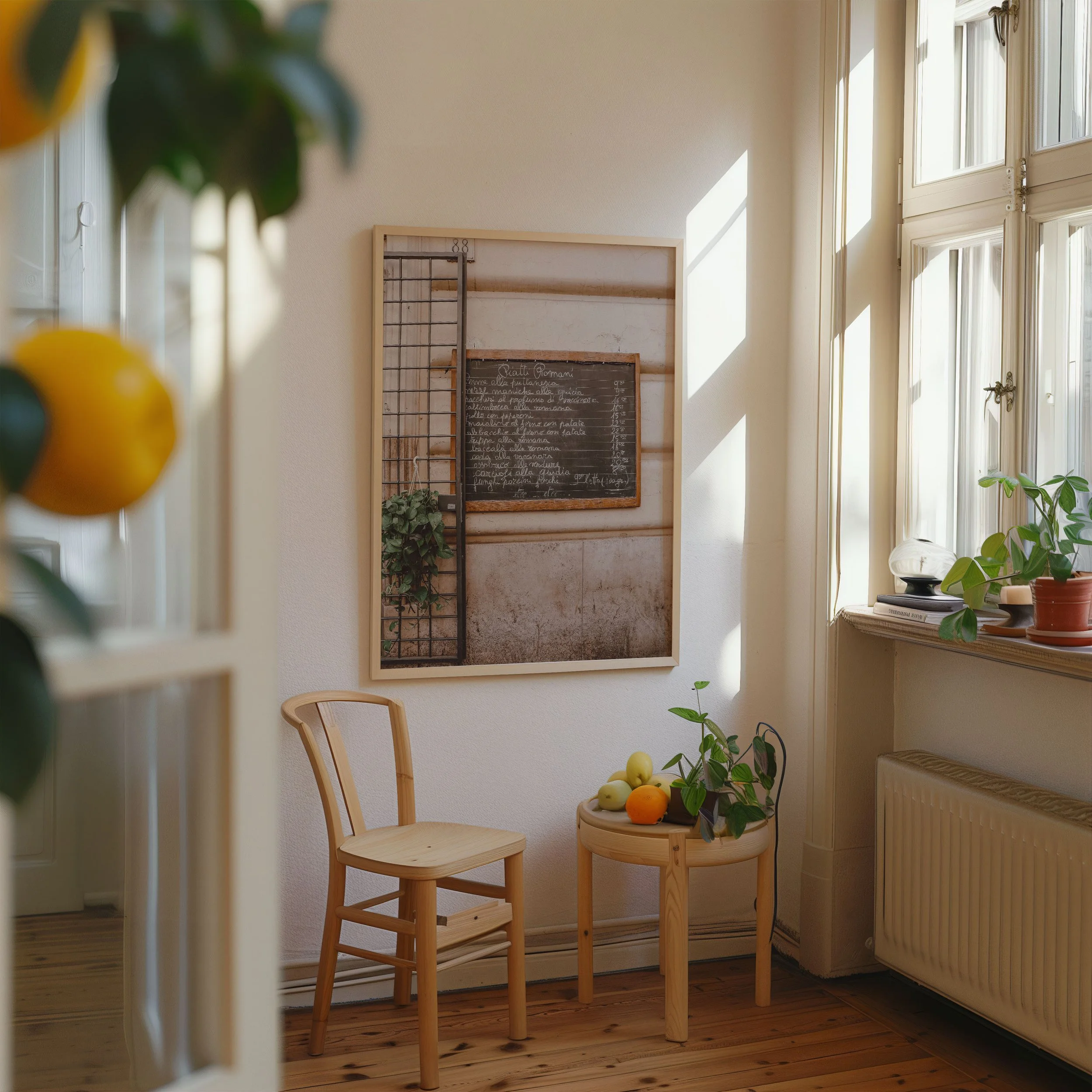 Cozy corner with wooden chair, small round table holding apples and greenery, large window with potted plant, and framed chalkboard on wall in a bright room with wooden floor.