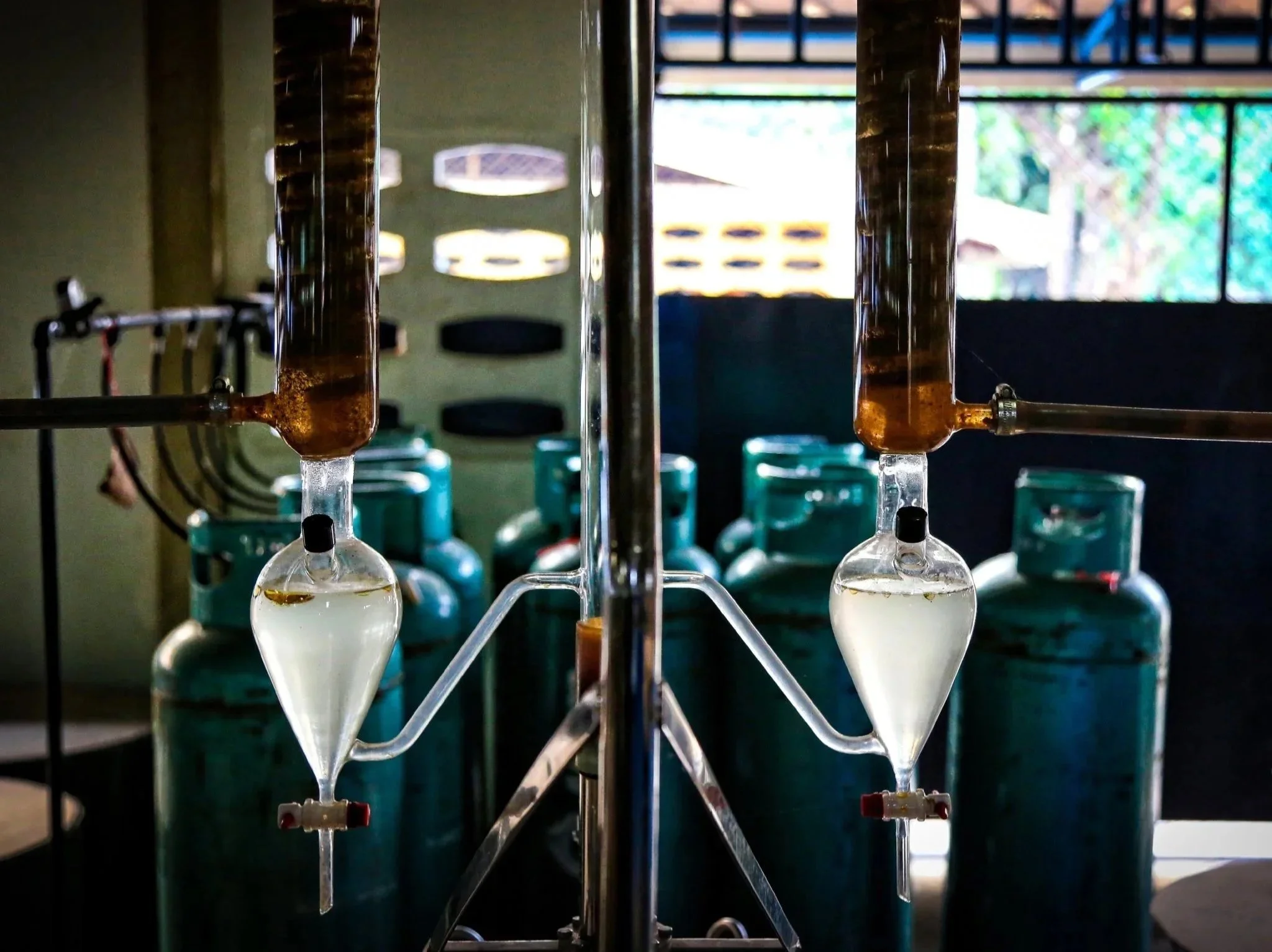 Laboratory setup with two separating funnels connected to a glass stand, with gas cylinders in the background, likely used for chemical experiments involving gases.