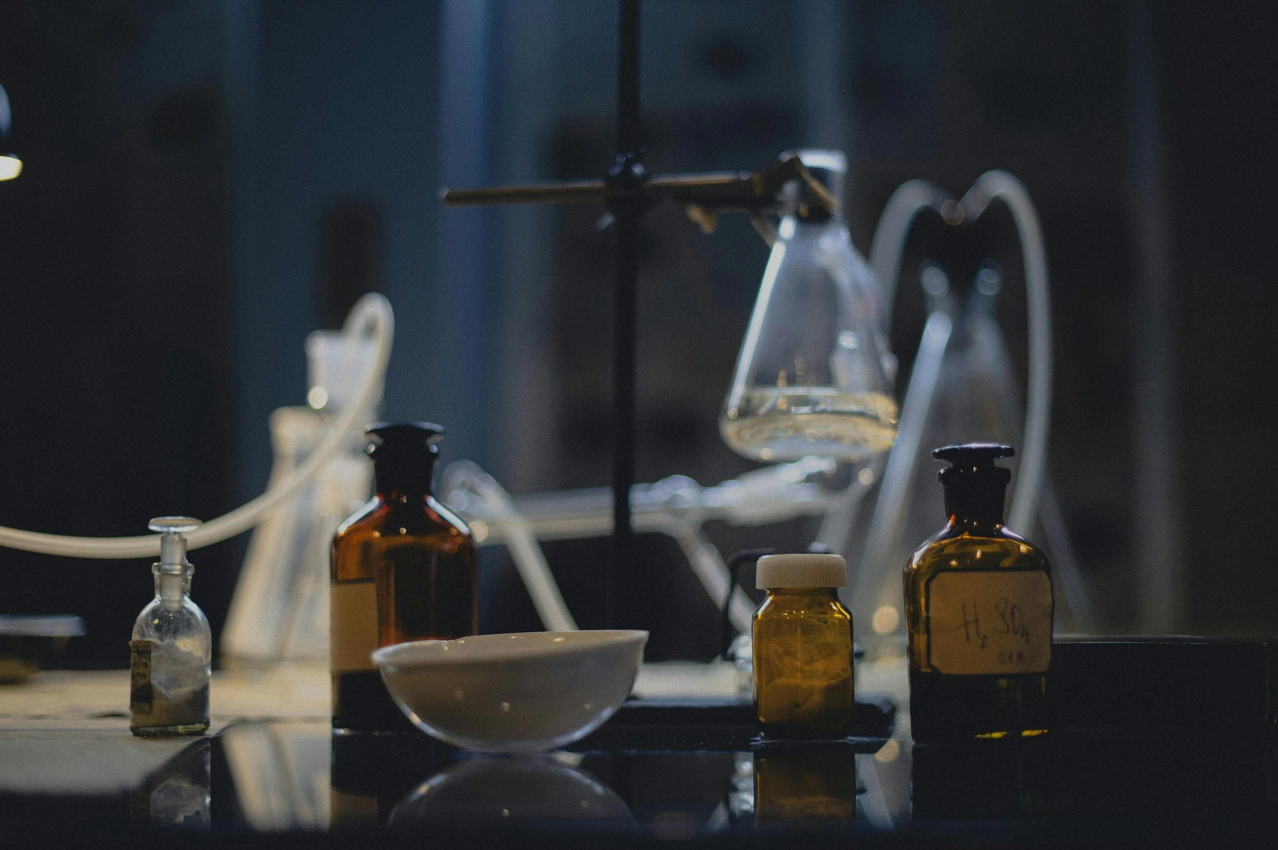 Laboratory bench with glassware and chemical bottles, with a glass container hanging from a stand and connected to tubes, in a dimly lit setting.