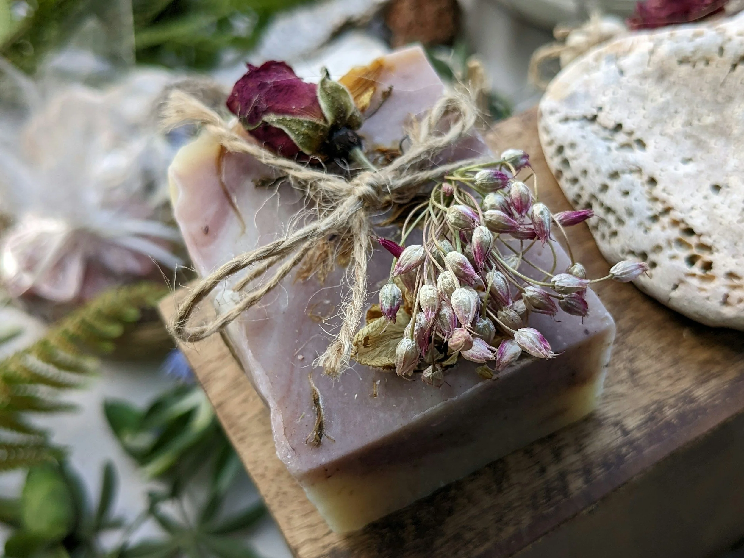 A bar of soap decorated with a bundle of dried flowers tied with twine, placed on a wooden surface next to a white, textured pumice stone.