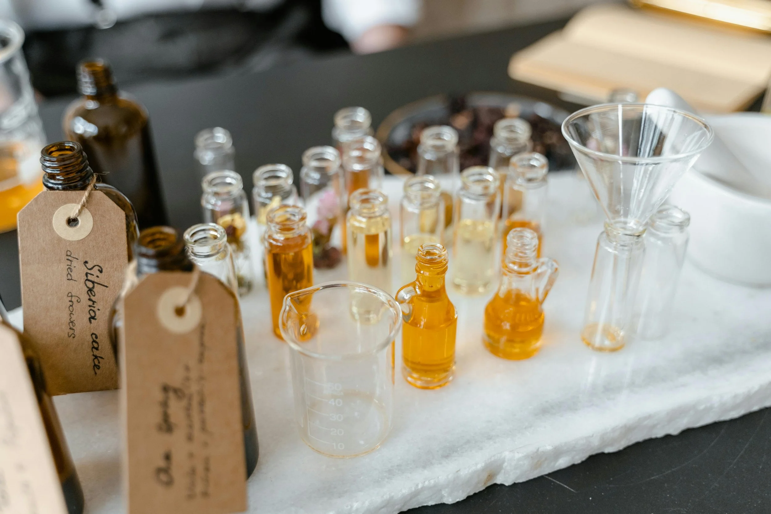 Various small glass bottles and jars filled with liquids, some labeled with tags, along with a beaker, a small funnel, and a bowl, arranged on a white textured surface.