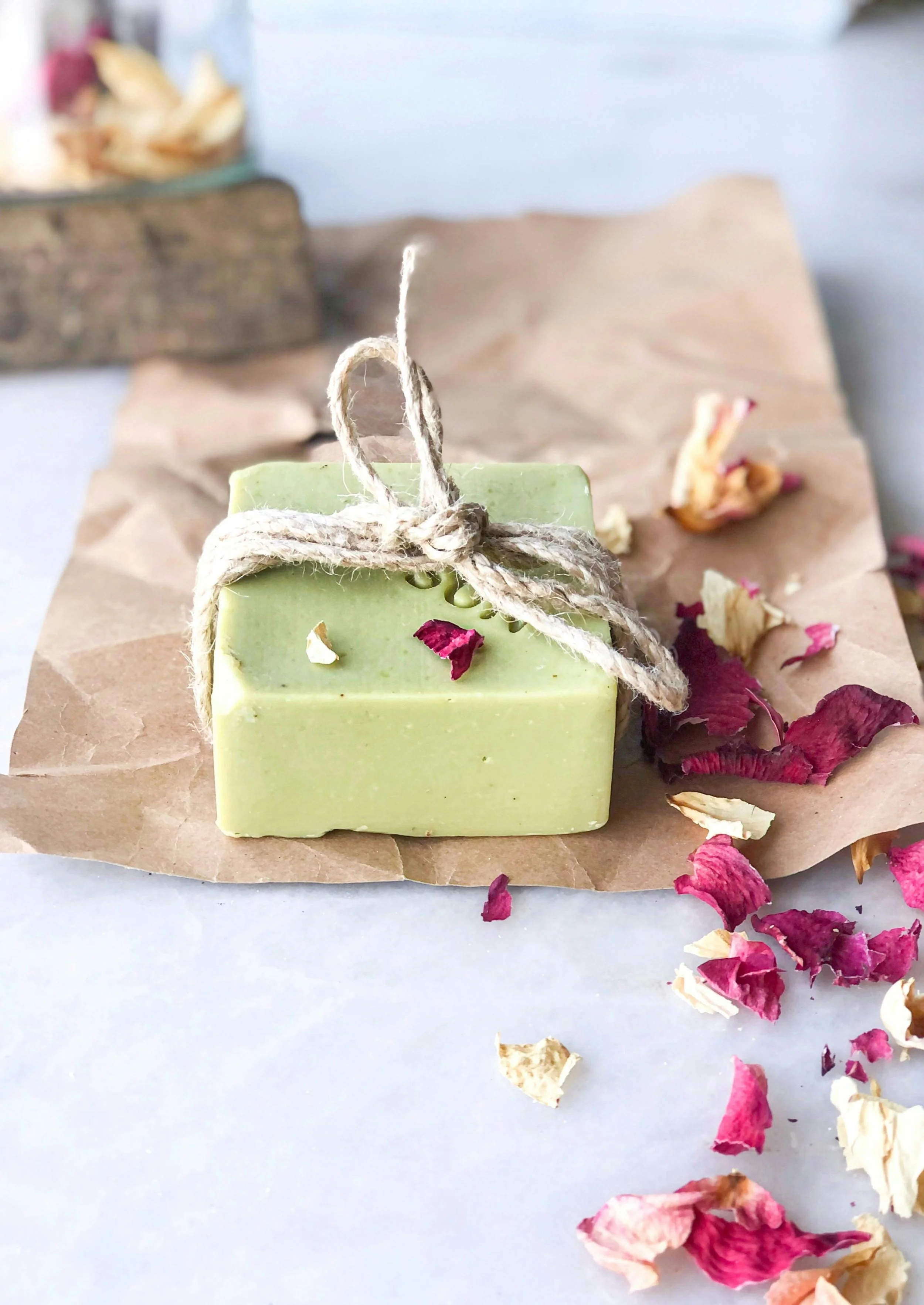 A green bar of soap tied with a jute string, placed on crumpled brown paper with dried rose petals scattered around.