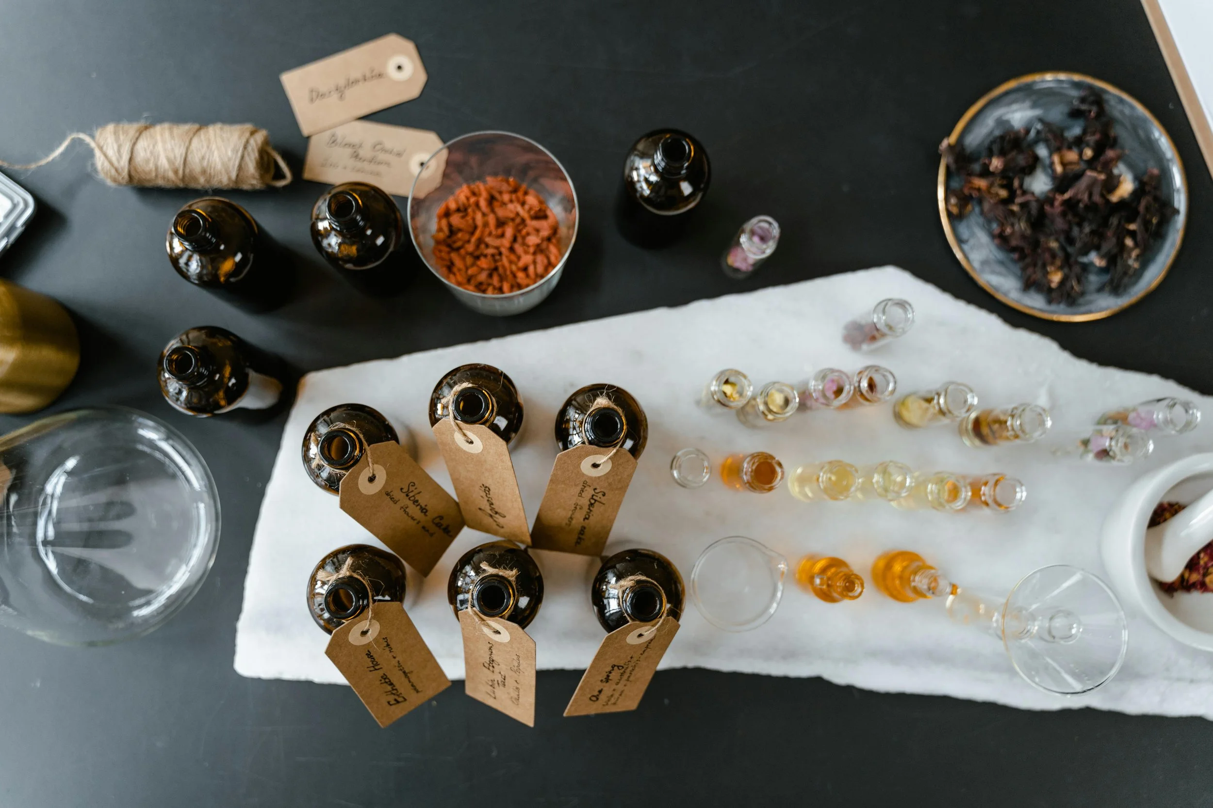 Top-down view of a table with various bottles and containers of essential oils, labeled tags, small glass vials, dried herbs, and a tea cup, set on a white cloth.