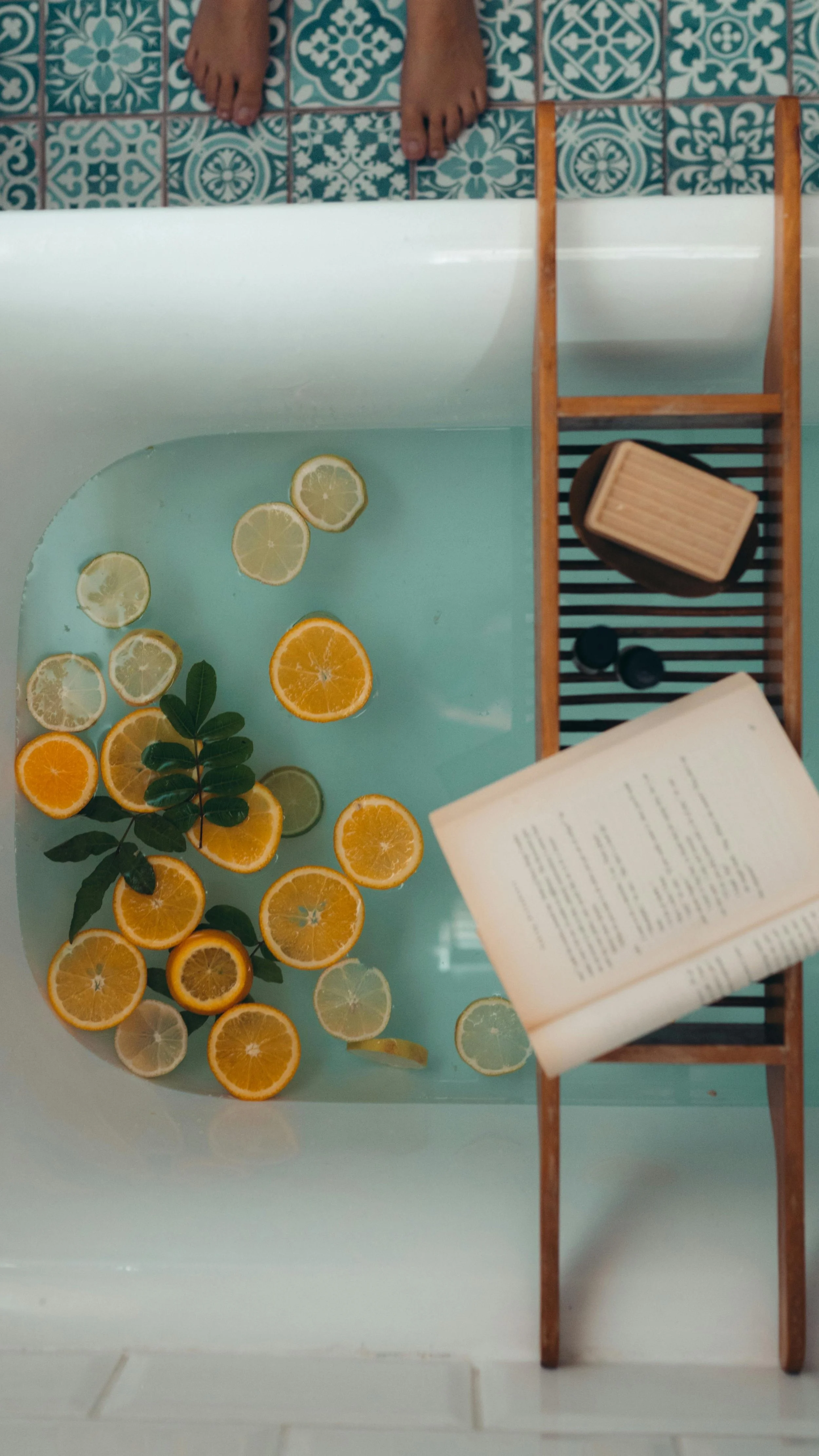 A bathtub filled with floating lemon and orange slices, with a book and some containers placed on a wooden bath caddy, viewed from above. The person's feet are visible at the top of the image, standing on patterned blue and white tiles.