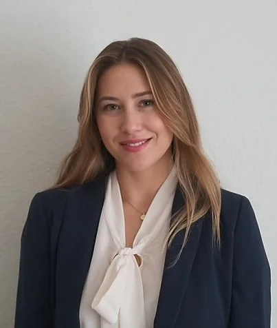 A young woman with blonde hair wearing a navy blazer and cream-colored blouse with a bow, standing against a plain white wall, smiling at the camera.