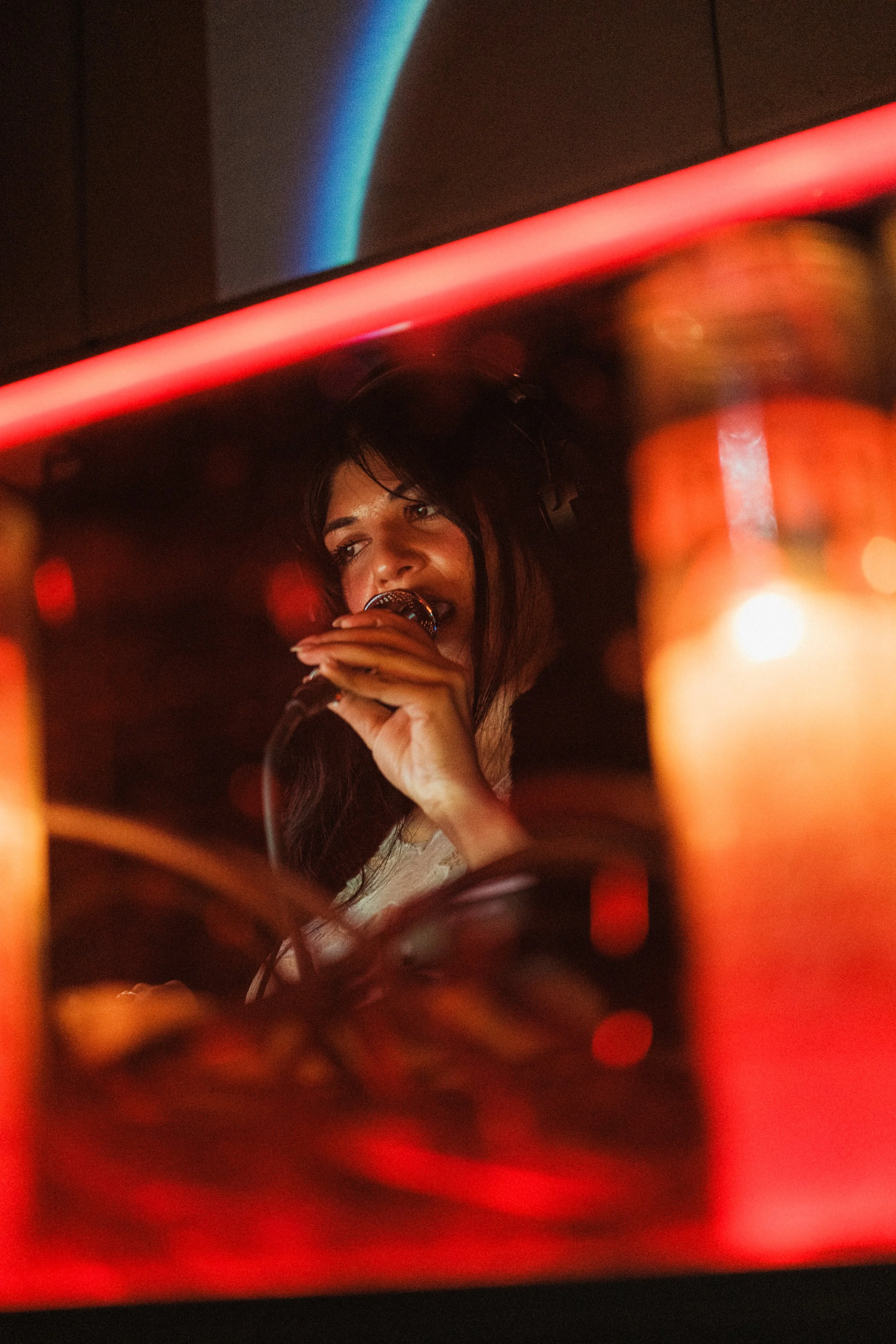 A woman singing into a microphone, seen through a reflective surface with red and blue lights.