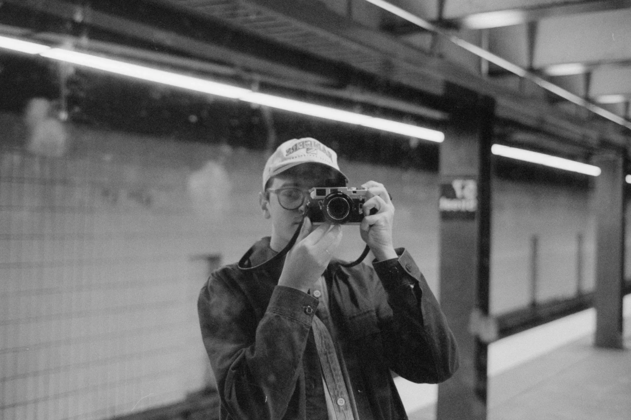 A person wearing glasses and a cap taking a photo with a camera in a subway station.