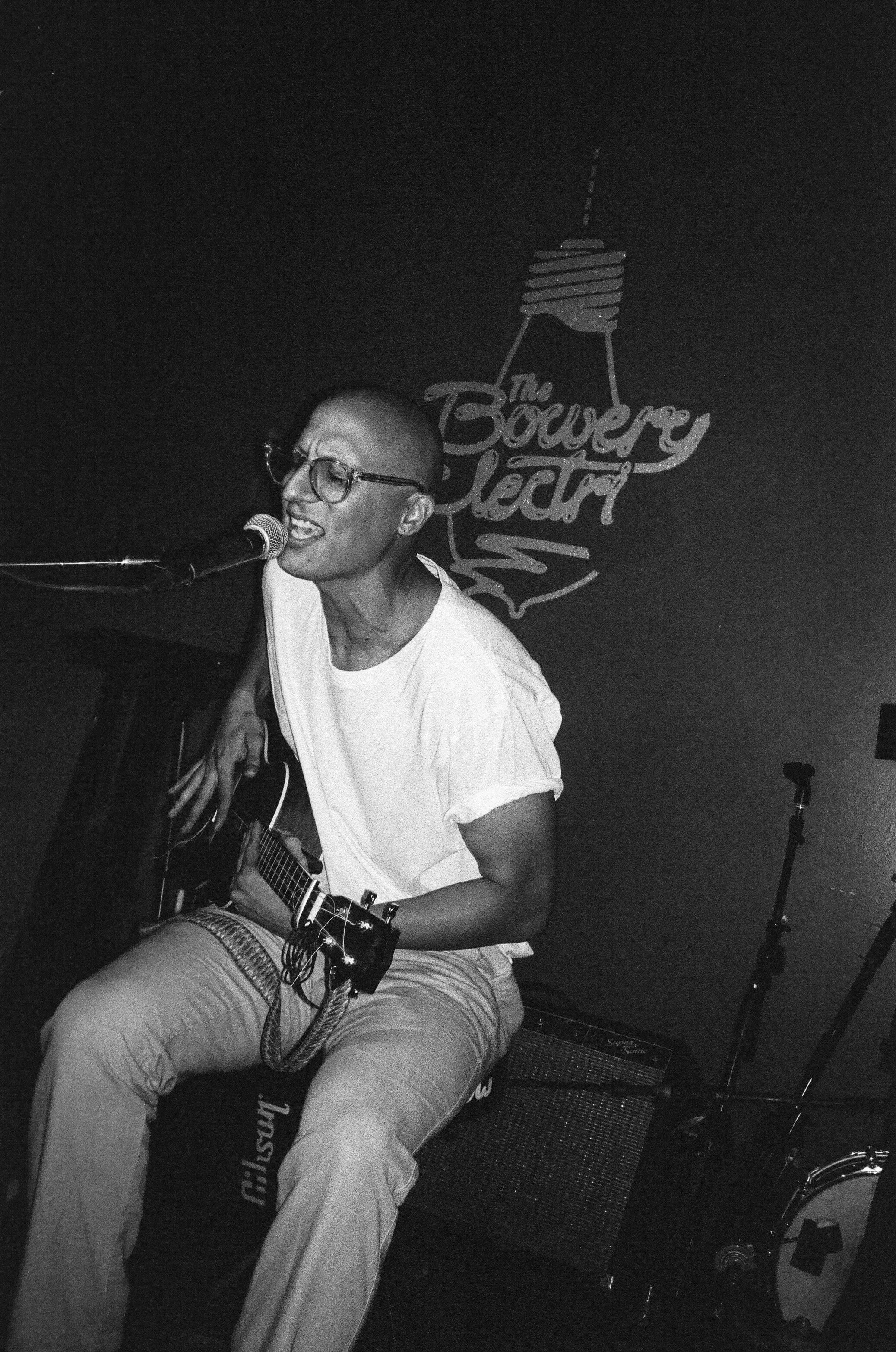 A person with glasses playing an acoustic guitar and singing into a microphone.