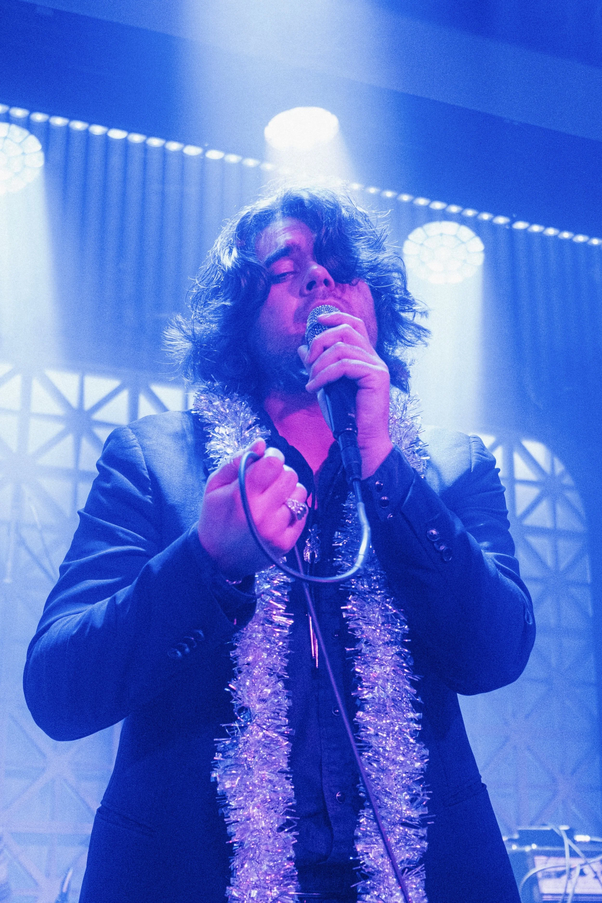 A male singer with curly hair singing into a microphone on stage decorated with a tinsel garland, under blue stage lighting.