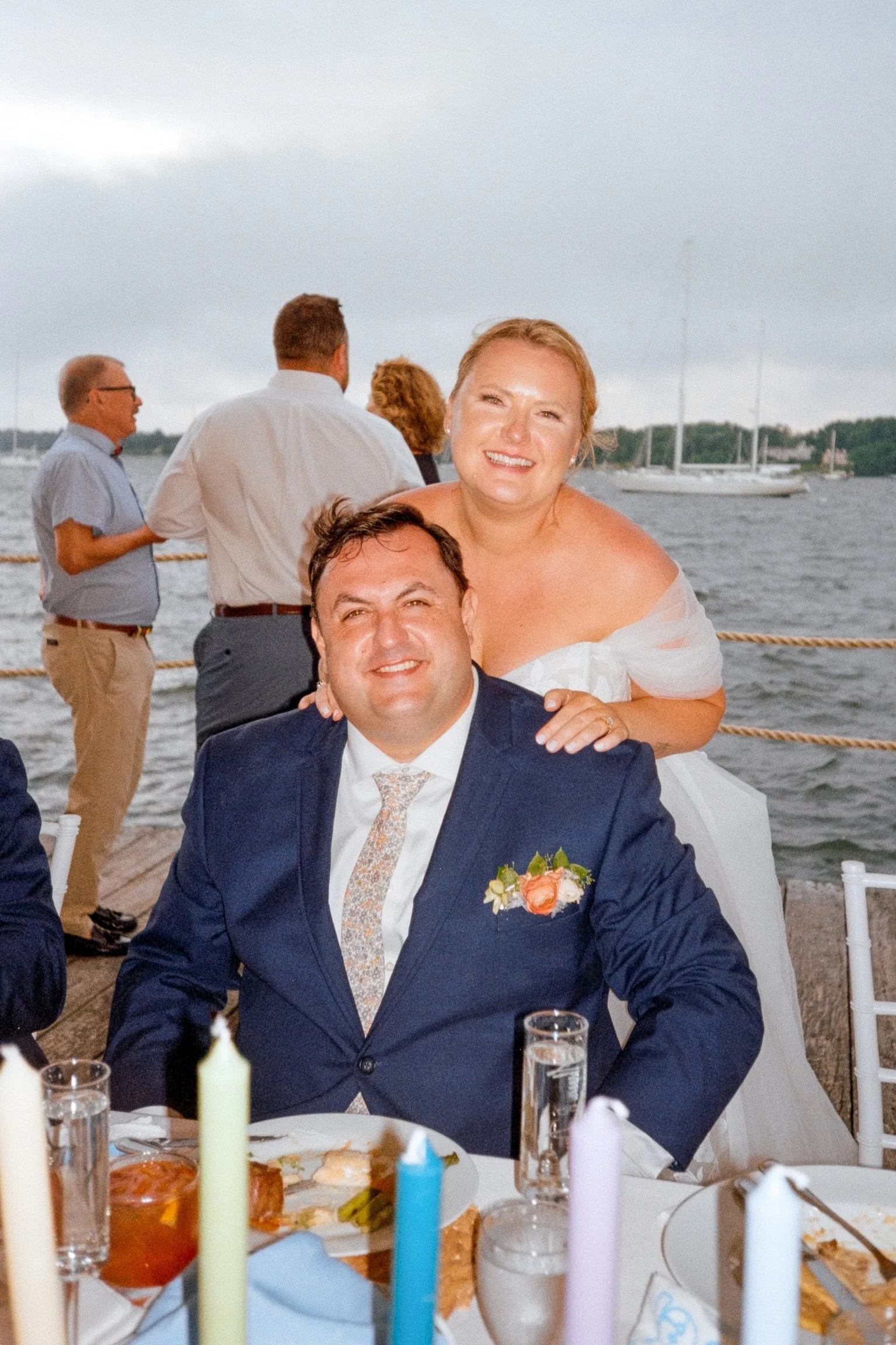 A smiling woman in a wedding dress stands behind a man in a navy suit with a floral tie, seated at a table outdoors near a body of water..