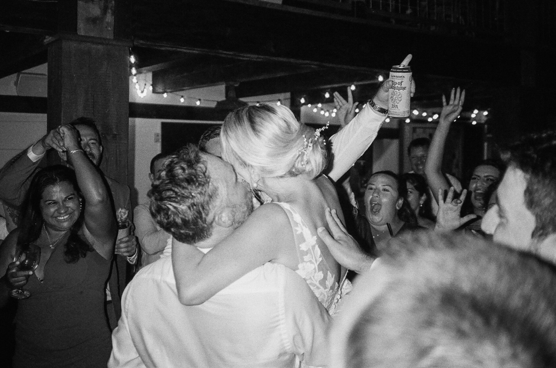Black and white photo of a wedding celebration with a couple kissing as guests cheer around them. One woman is holding a drink and raising her hands, and others are smiling and dancing.