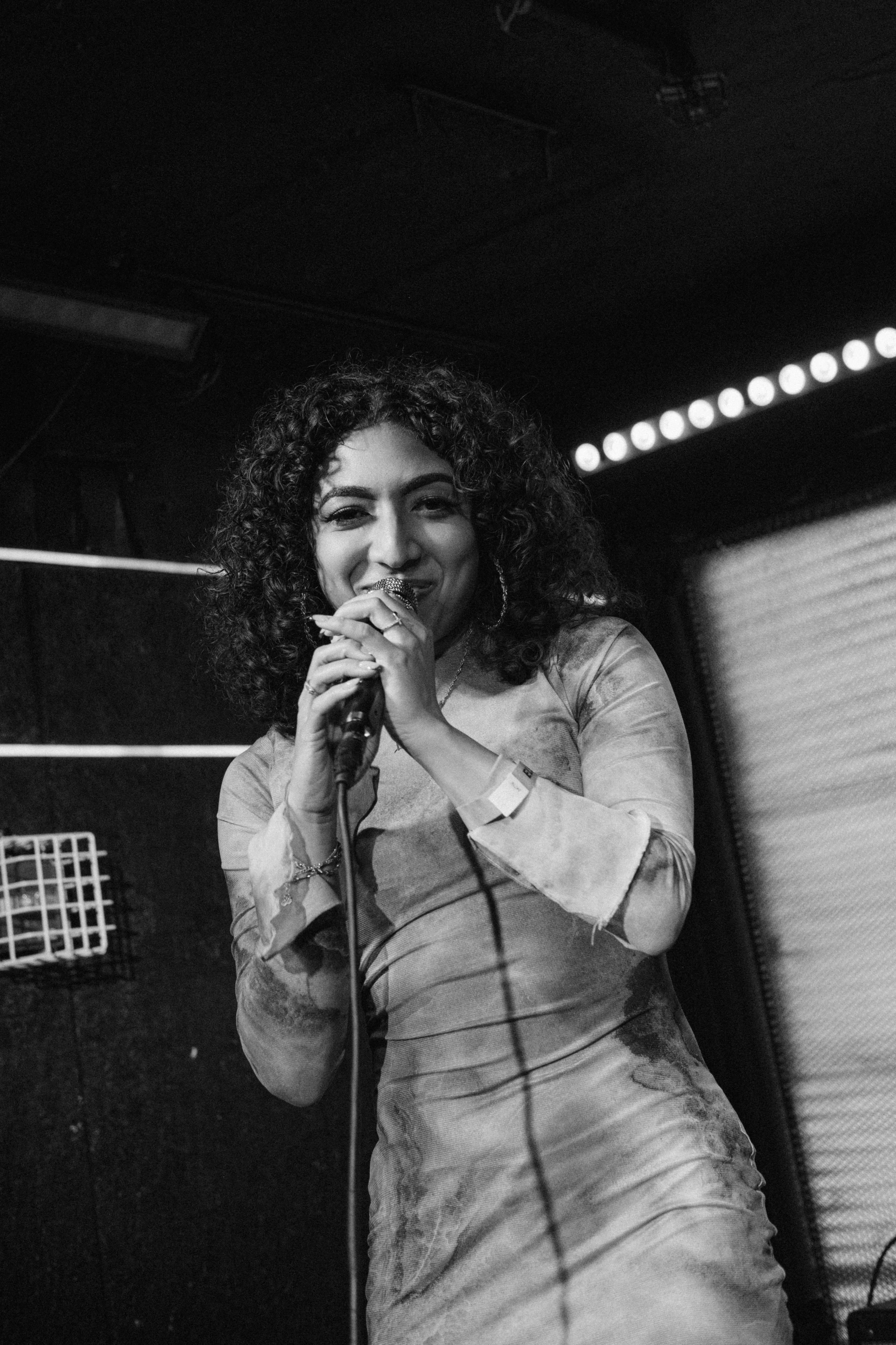 Black and white photo of a woman singing into a microphone on stage, with a smile, wearing a fitted dress and hoop earrings.