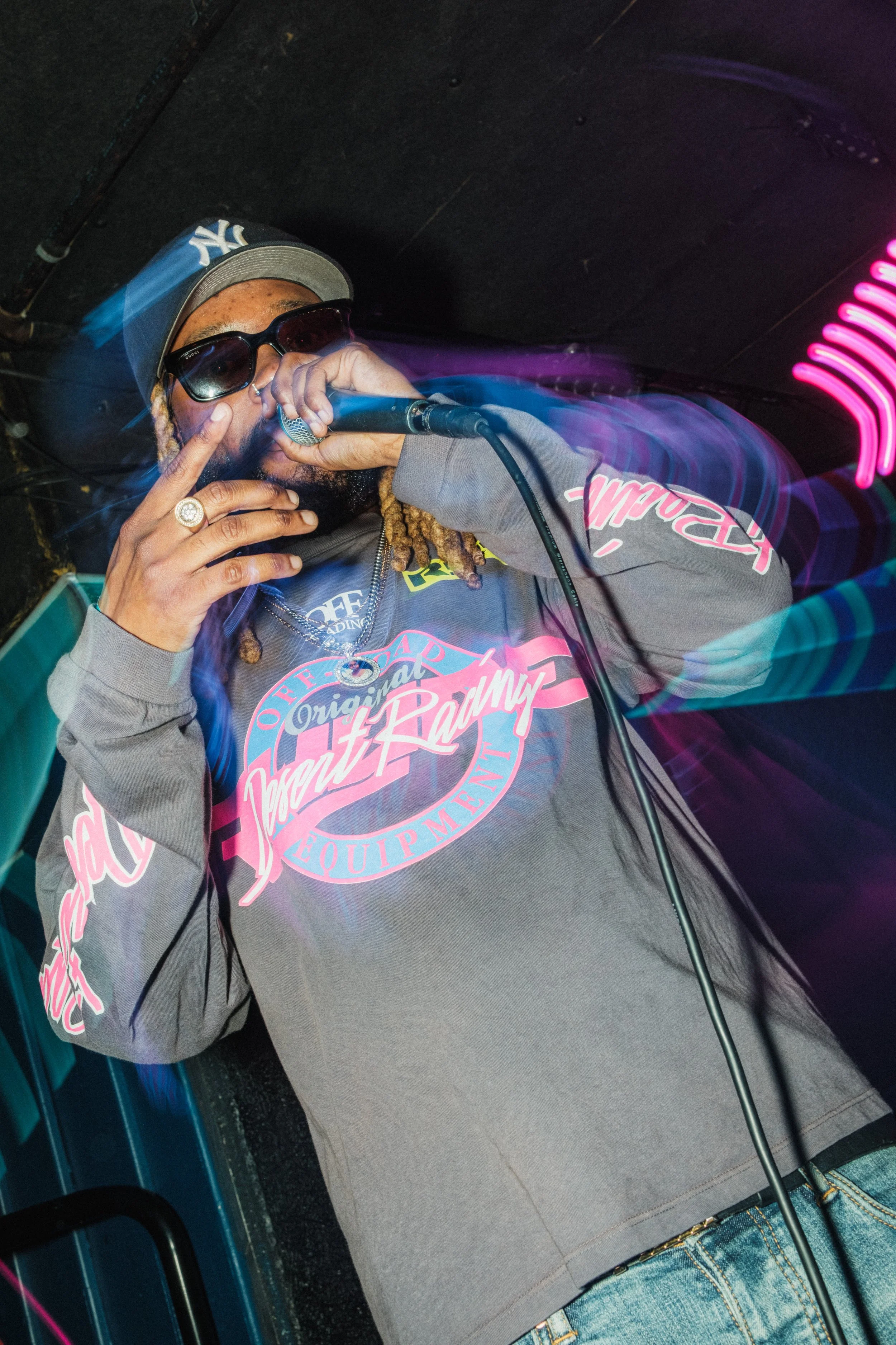 A rapper with dreadlocks wearing a gray hoodie with pink and blue graphics, sunglasses, and a baseball cap, performing on stage.