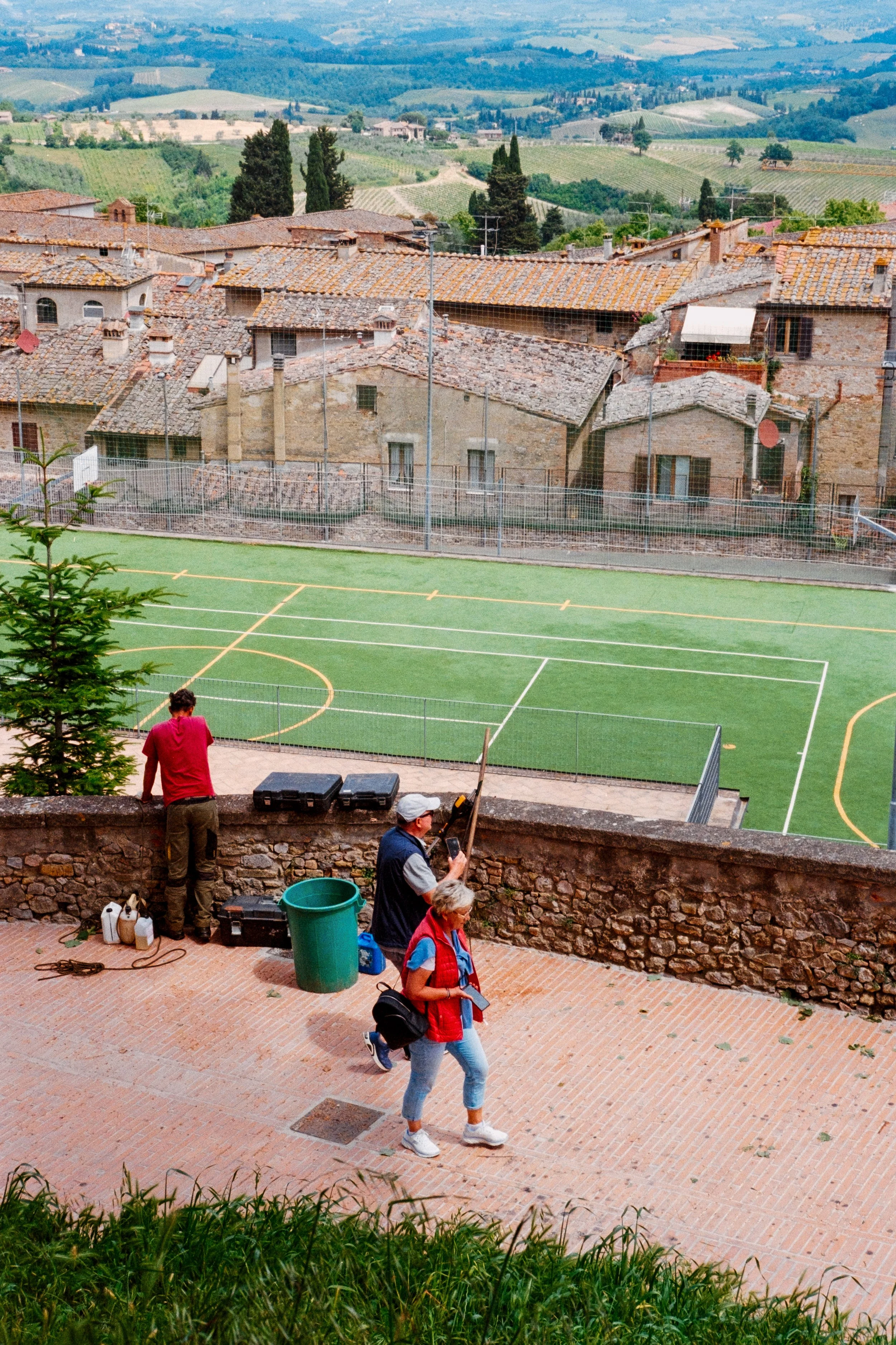 A view from high up in San Gimignano, Tuscany, with people walking in the foreground, a small soccer pitch in the middle ground, and the rolling countryside in the background.