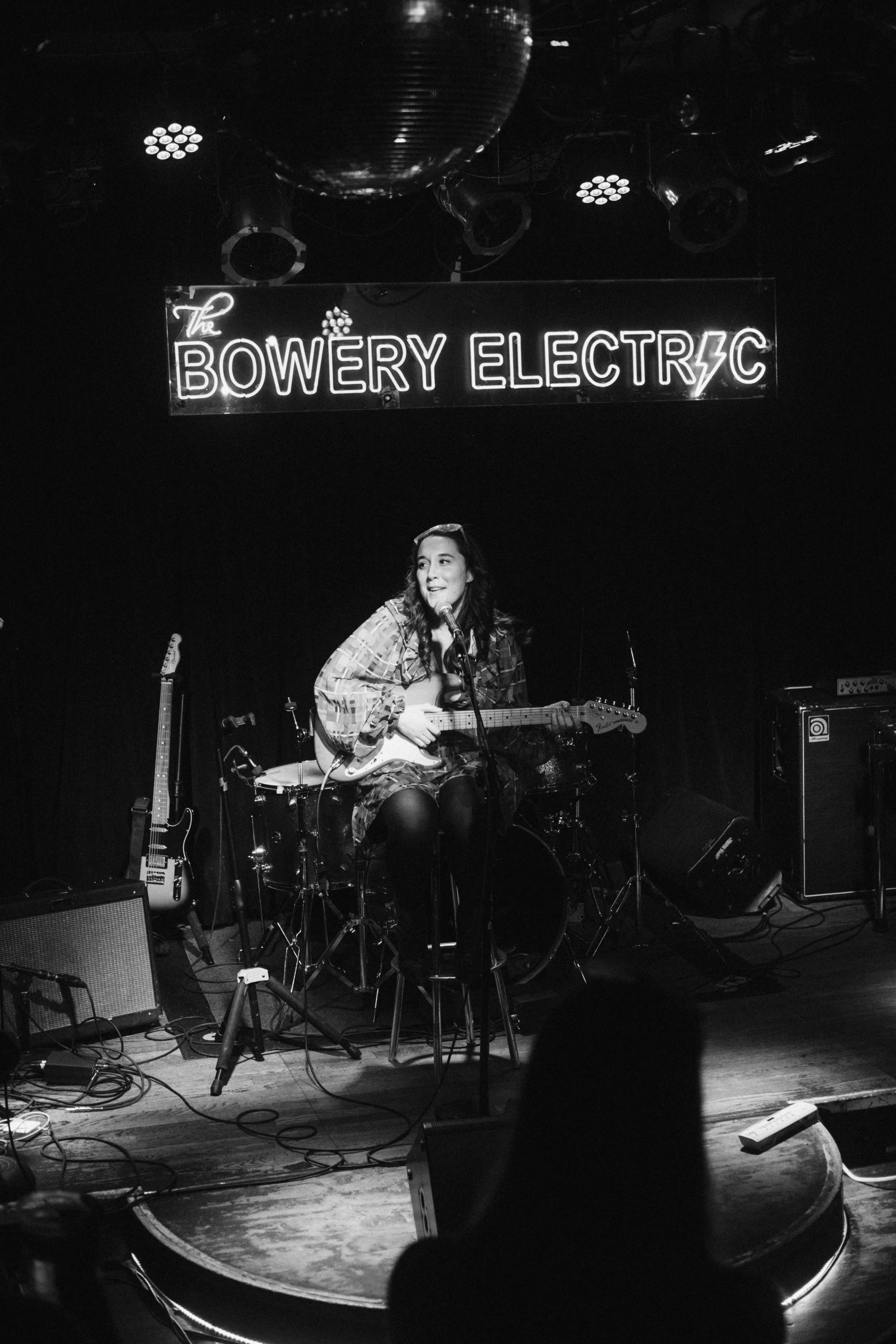 A female musician sitting on a stool with a guitar, performing on stage at The Bowery Electric.