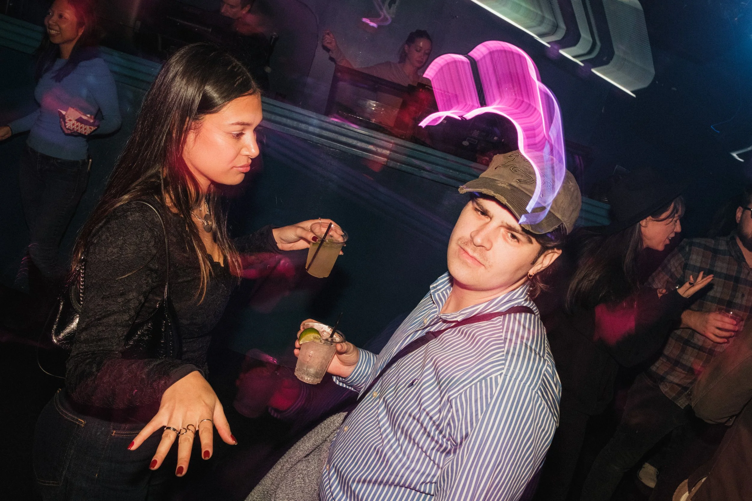 People socializing at a nightclub, holding drinks with colorful lighting and neon decorations in the background.