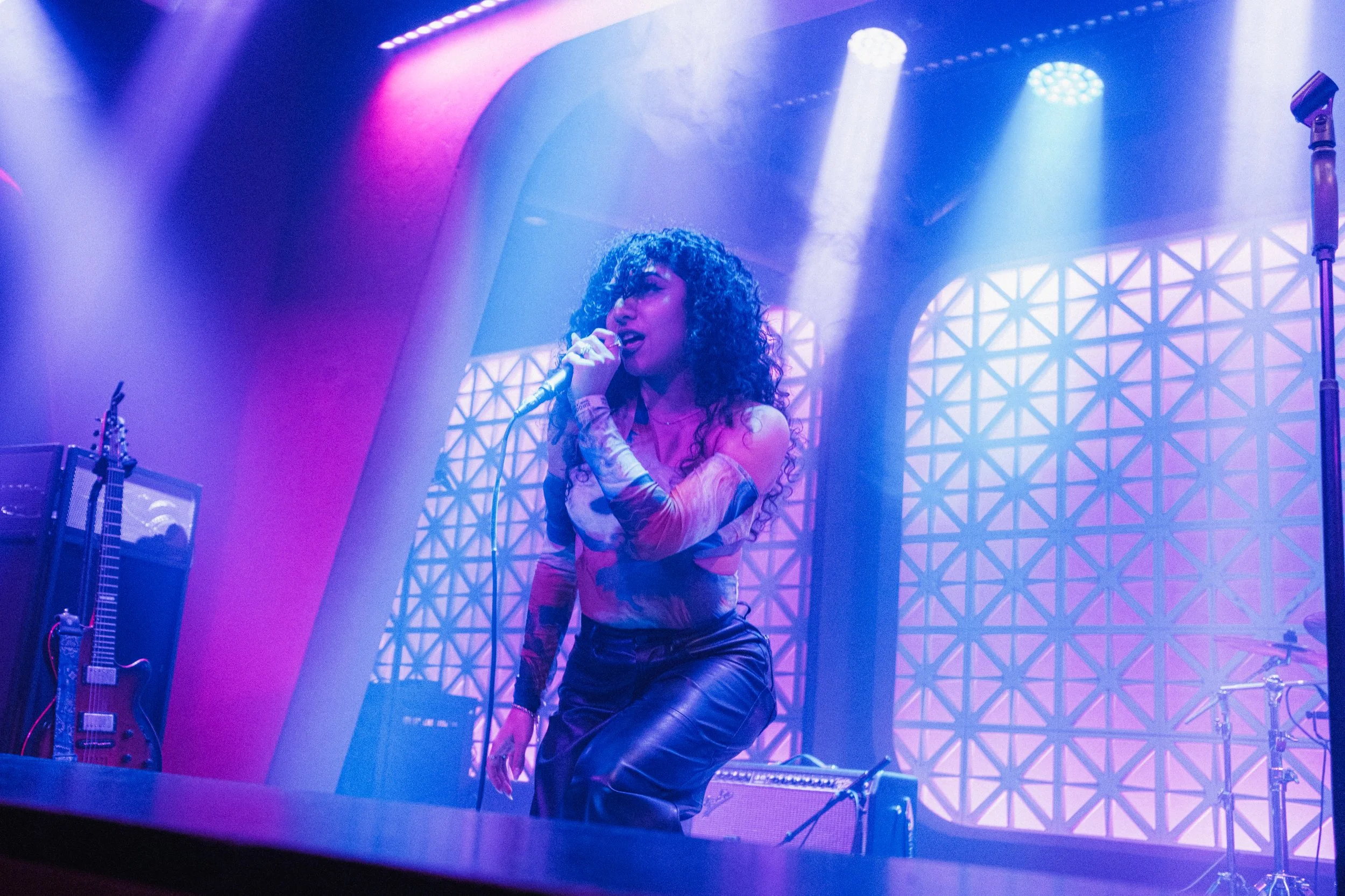 A female singer with curly hair performing on stage, holding a microphone, with colorful stage lights overhead.