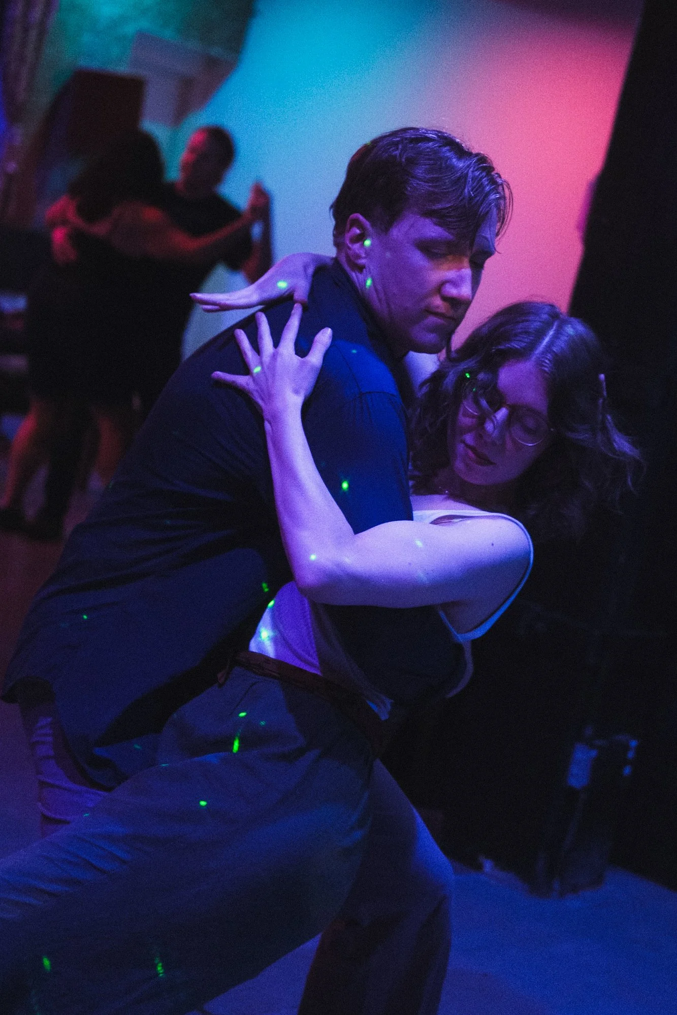 A couple dancing closely together in a dimly lit nightclub with colorful lights, with other dancers in the background.