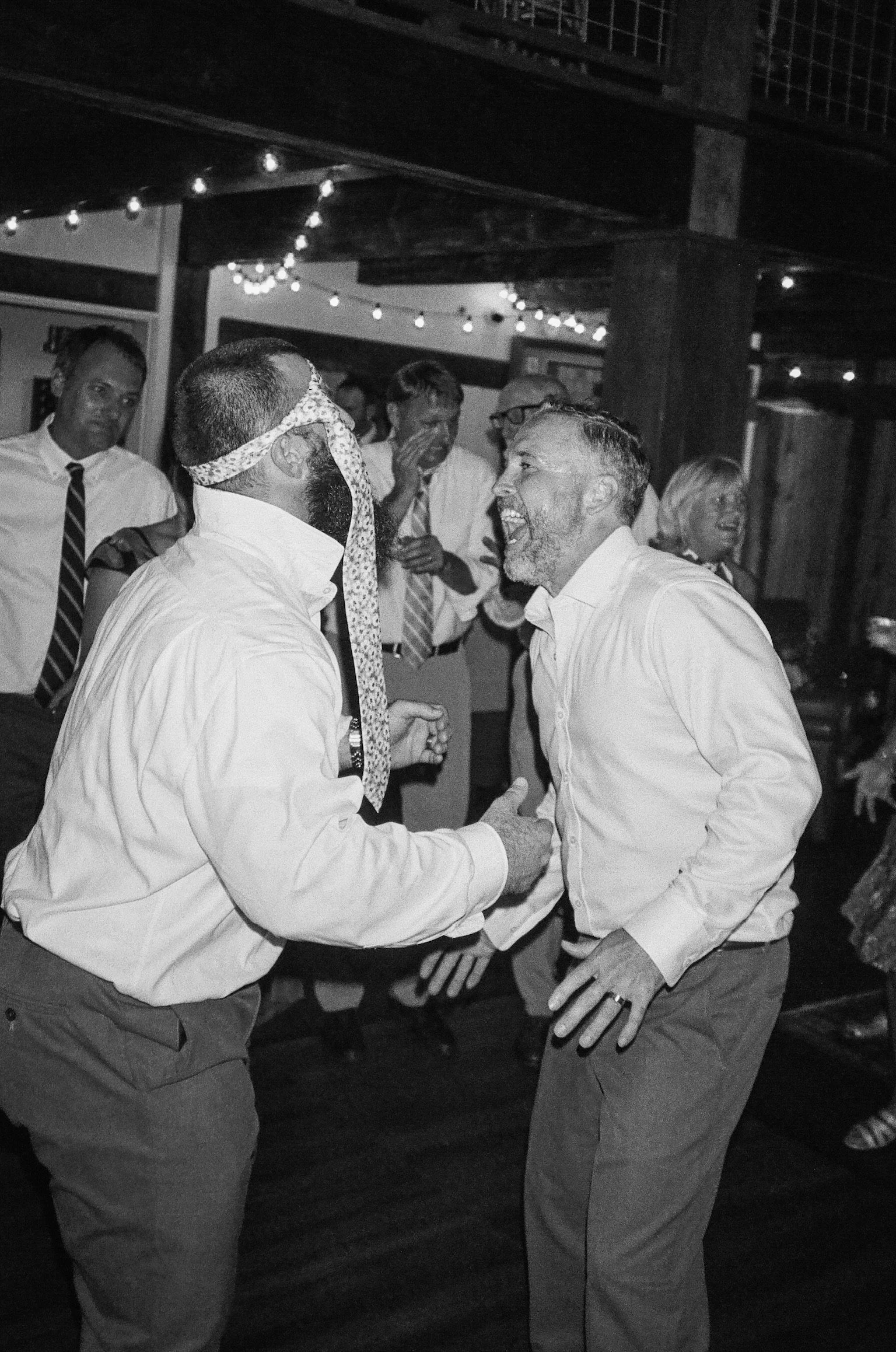 People dancing and celebrating at a wedding reception, with one person blindfolded with a necktie and holding hands with another.