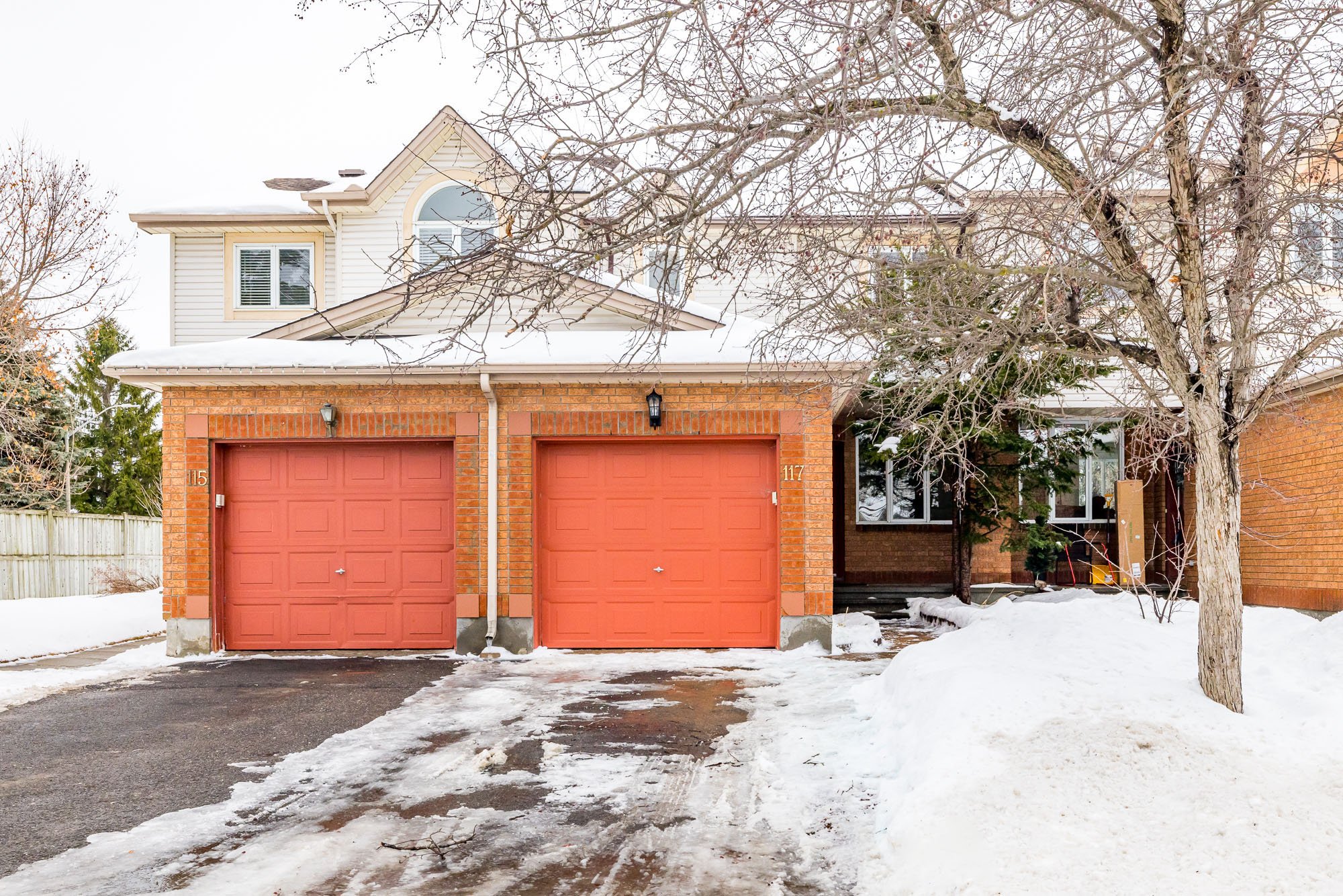 Front view of a house with two red garage doors, brick exterior, and snow on the ground, with a leafless tree in the foreground.